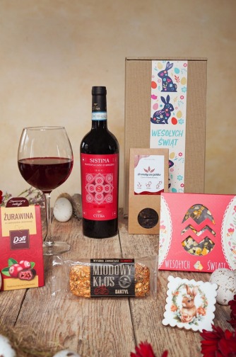Zestaw prezentowy na Wielkanoc: wino Sistina, miód daktilowy, czekoladki żurawinowe, herbata i dekoracje w pudełku z motywem zajączków na drewnianym stole.