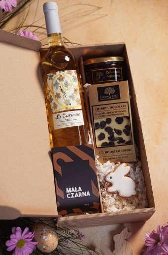 Elegancki prezent w pudełku: butelka białego wina La Curieuse, konfitura wiśniowa, czekolada Cherry Tree i ciasteczko w kształcie zajączka, udekorowane kwiatami i pisanką.