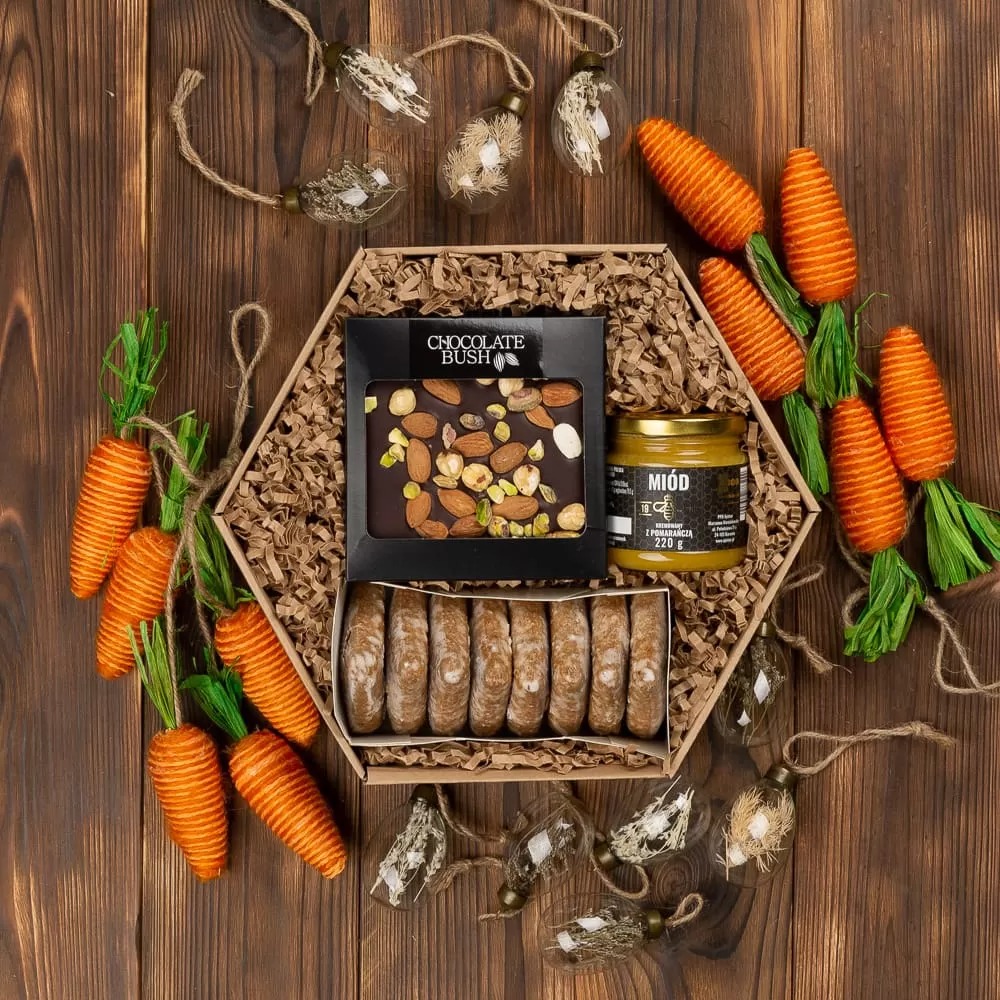 Ozdobny, heksagonalny prezentowy box z czekoladą Chocolate Bush z orzechami, miodem pomarańczowym 220g, piernikami i dekoracjami w kształcie marchewek i szklanych bombek na sznurku, na tle...