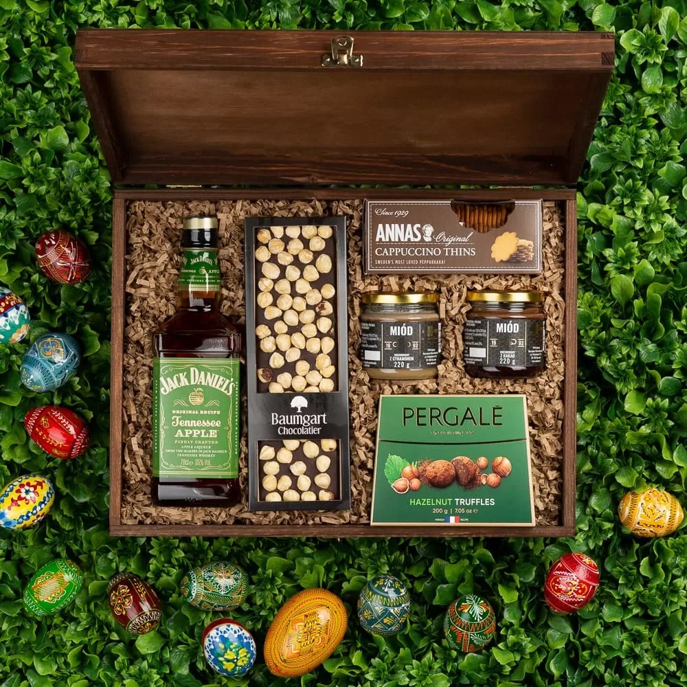 Drewniana skrzynka prezentowa wypełniona whisky, czekoladą z orzechami, miodem, ciasteczkami i truflami, otoczona pisankami na tle zieleni.