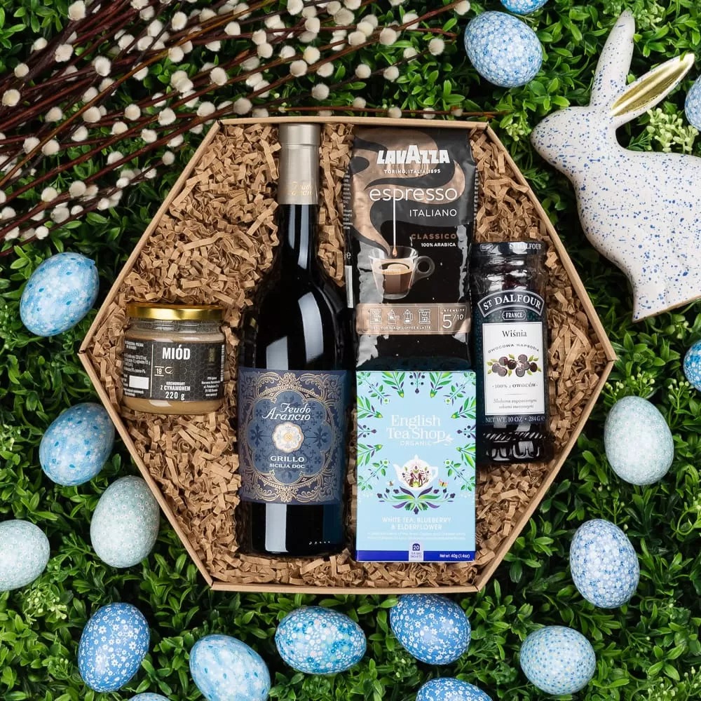 Heksagonalny kosz prezentowy wypełniony miodem, winem Grillo, kawą Lavazza Espresso Italiano, herbatą English Tea Shop i konfiturą wiśniową St Dalfour, otoczony dekoracyjnymi, niebieskimi jajkami...