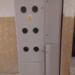 Szara szafa elektryczna z okrągłymi otworami na liczniki, umieszczona przy ścianie o wzorzystej fakturze.