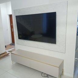 RAF-ART - Nowoczesny salon po remoncie: Telewizor na ścianie wykończonej marmurem, beżowa szafka RTV z złotymi akcentami, w tle elementy remontu - kartony i folie ochronne.