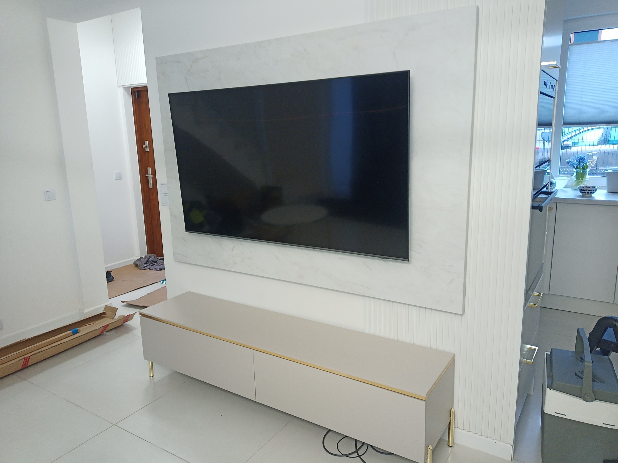 Nowoczesny salon po remoncie: Telewizor na ścianie wykończonej marmurem, beżowa szafka RTV z złotymi akcentami, w tle elementy remontu - kartony i folie ochronne.