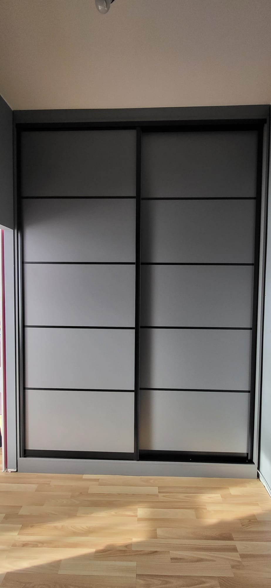 Szara szafa wnękowa z przesuwnymi drzwiami, podzielonymi na poziome panele, na drewnianej podłodze.