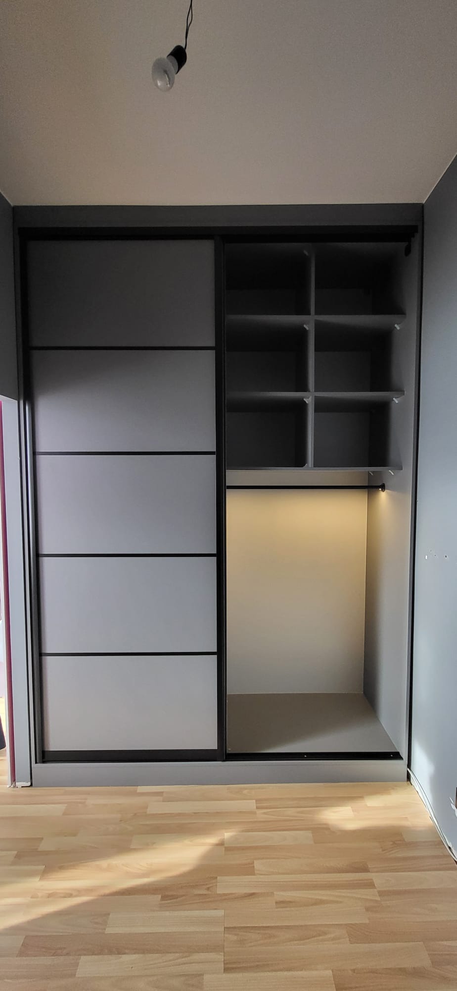 Szara szafa wnękowa z przesuwnymi drzwiami i oświetleniem LED wewnątrz, widoczne półki i drążek na ubrania, na podłodze panele imitujące drewno.