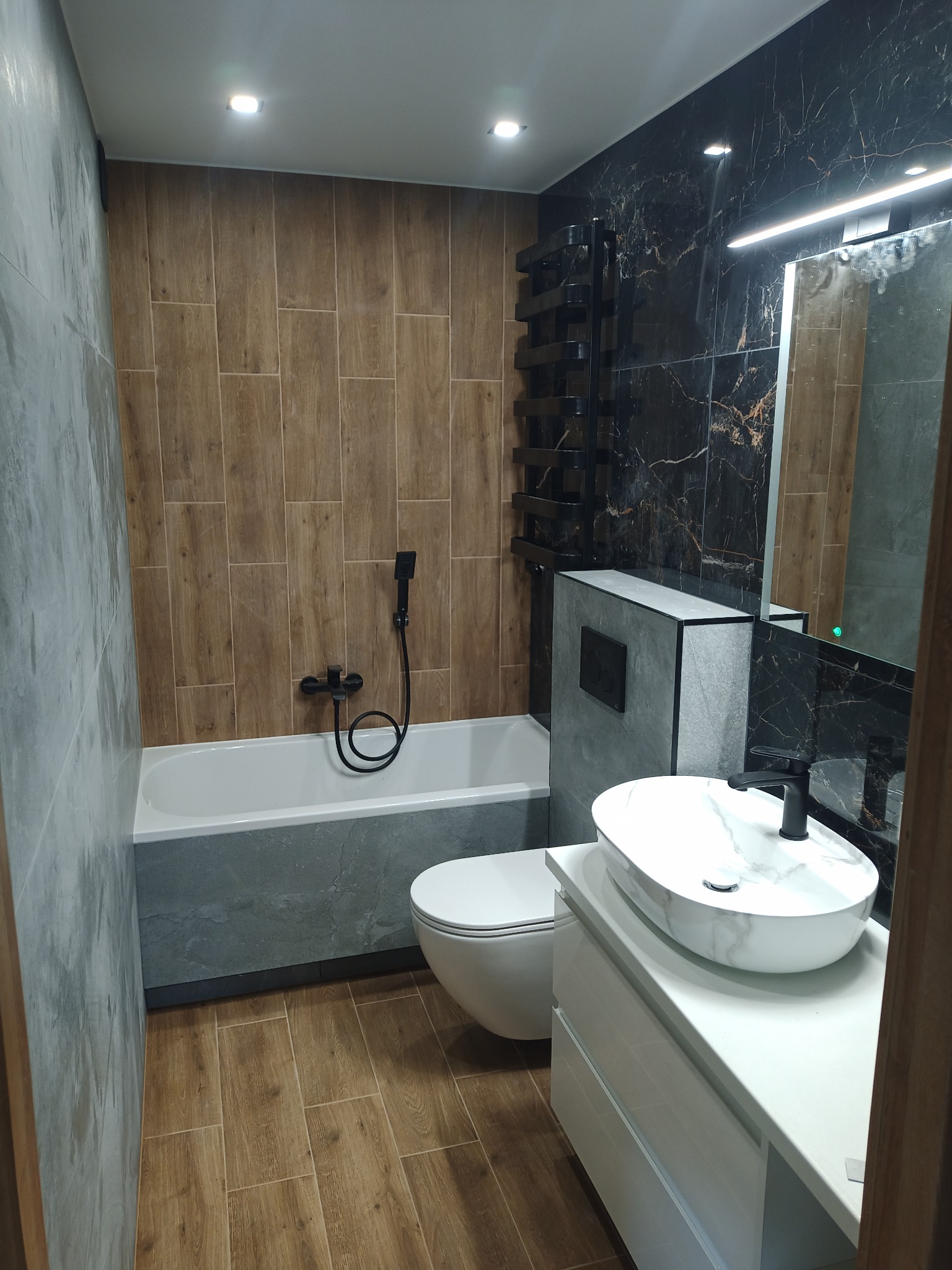 Nowoczesna łazienka z płytkami imitującymi drewno i marmur, czarna armatura, umywalka nablatowa na białej szafce, grzejnik drabinkowy.