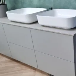 Dwie kwadratowe, białe umywalki nablatowe na szarej szafce z czterema frontami, na podłodze parkiet ułożony we wzór jodełki.