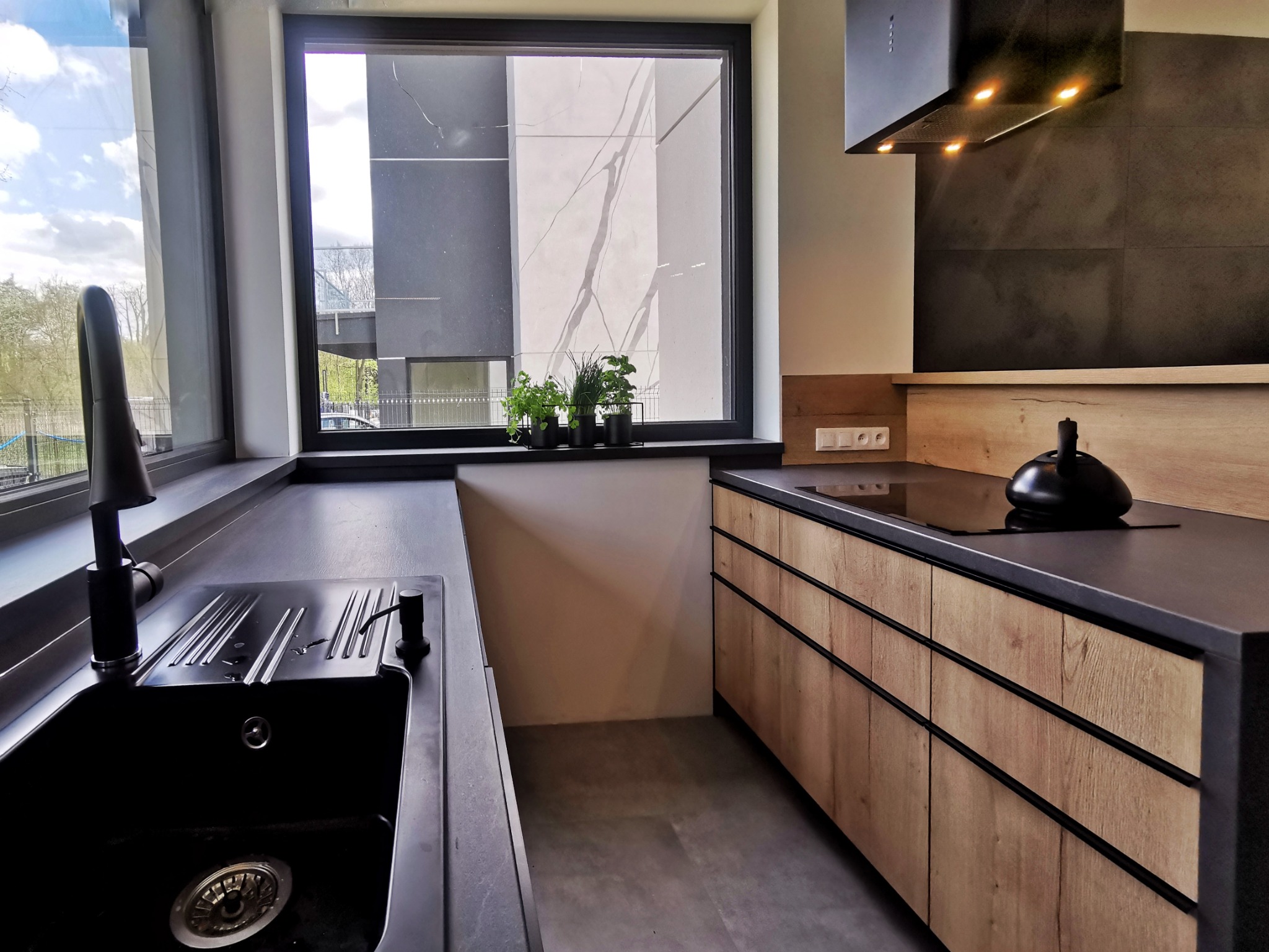Nowoczesna kuchnia z drewnianymi frontami szafek, czarnym blatem i zlewozmywakiem, oraz roślinami w doniczkach na parapecie okna.
