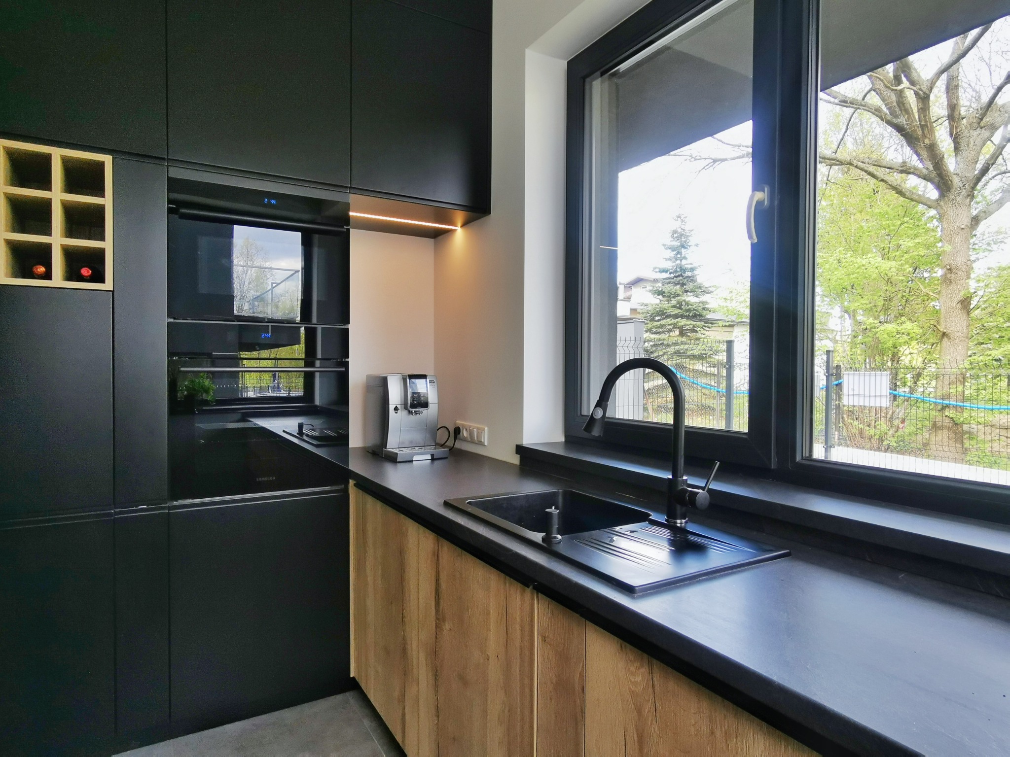 Nowoczesna kuchnia z czarnymi matowymi szafkami, drewnianymi frontami dolnymi i czarnym zlewem pod oknem z widokiem na zieleń.