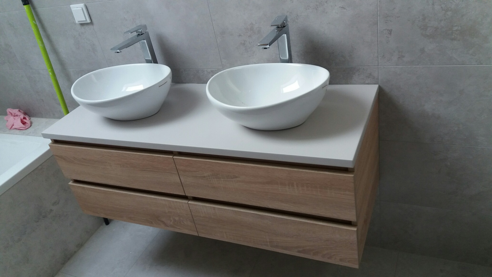 Nowoczesna łazienka z podwójną umywalką nablatową, drewniana szafka z czterema szufladami, szare płytki na ścianie, minimalistyczne baterie.