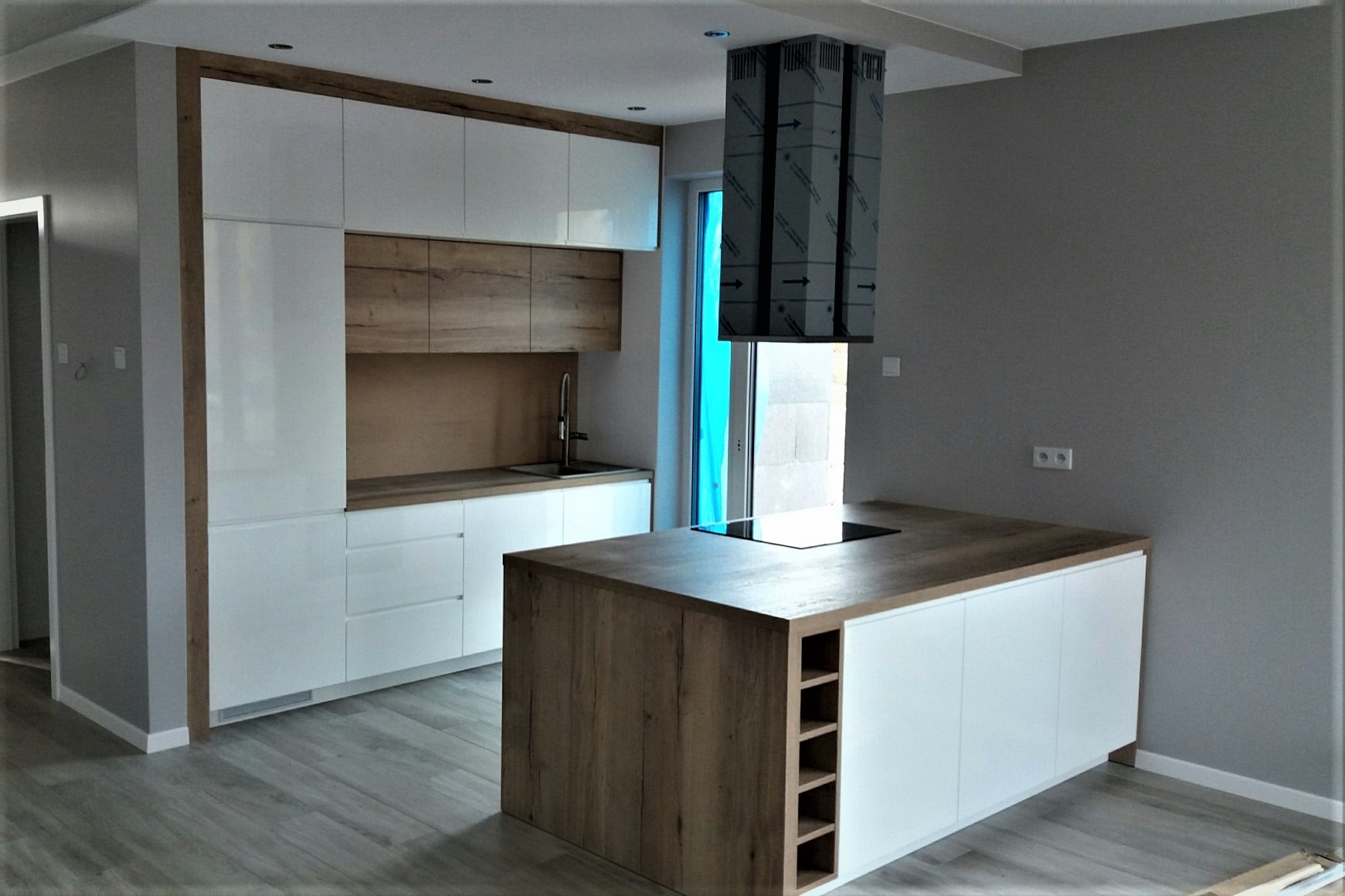 Nowoczesna kuchnia z białymi szafkami i drewnianymi akcentami, wyspa kuchenna z płytą indukcyjną i półkami na wino, sufitowy okap w folii ochronnej, szare ściany i jasna podłoga.