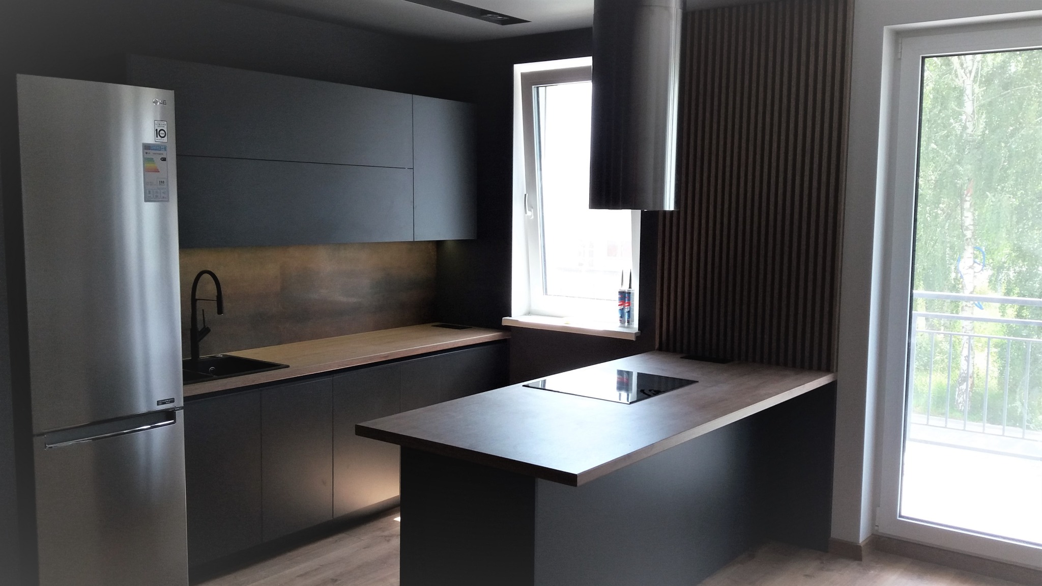 Nowoczesna kuchnia w ciemnej kolorystyce z wyspą, srebrna lodówka, drewniane blaty i pionowe panele dekoracyjne na ścianie.