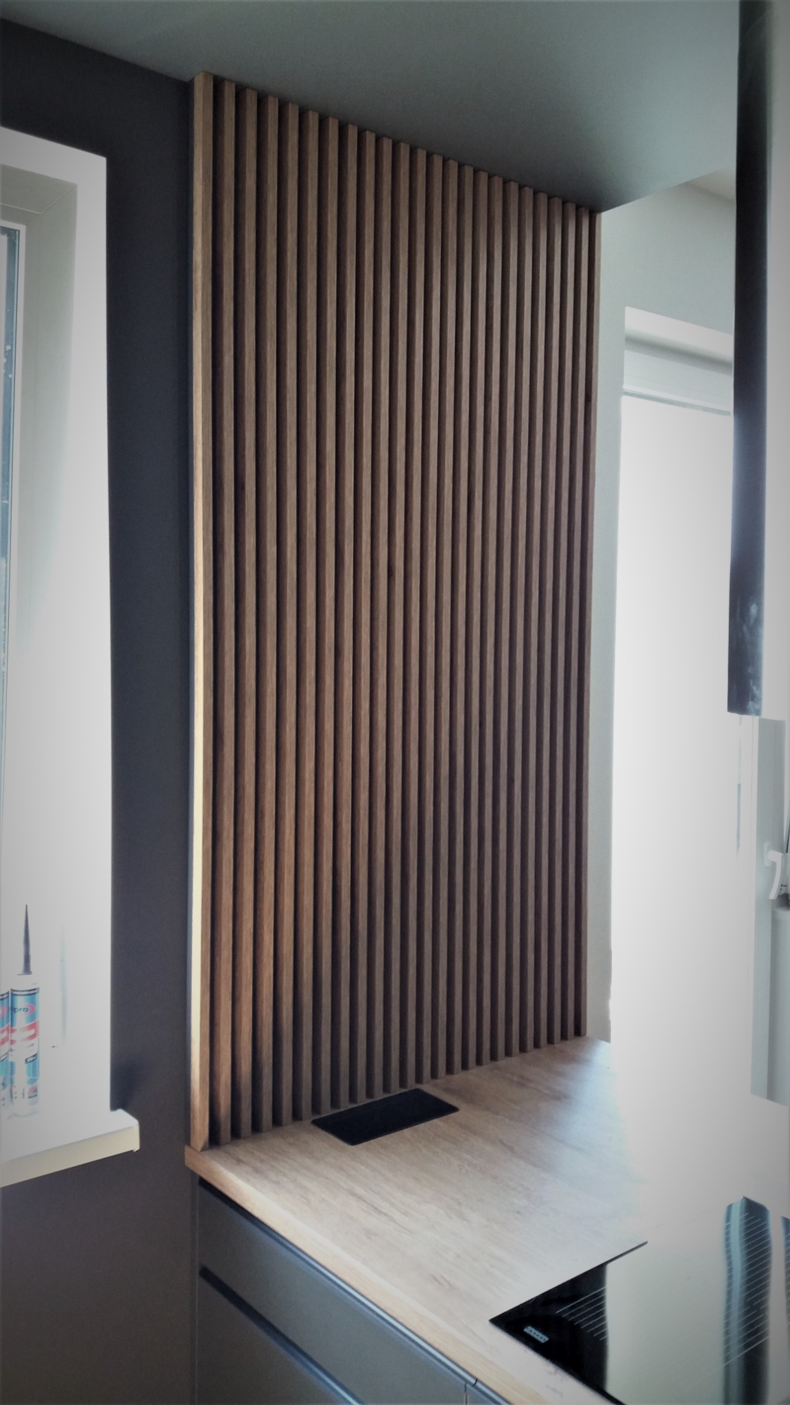 Drewniana ścianka działowa z pionowych listew w kuchni, blat z gniazdem elektrycznym, szare ściany i okno z akcesoriami montażowymi.