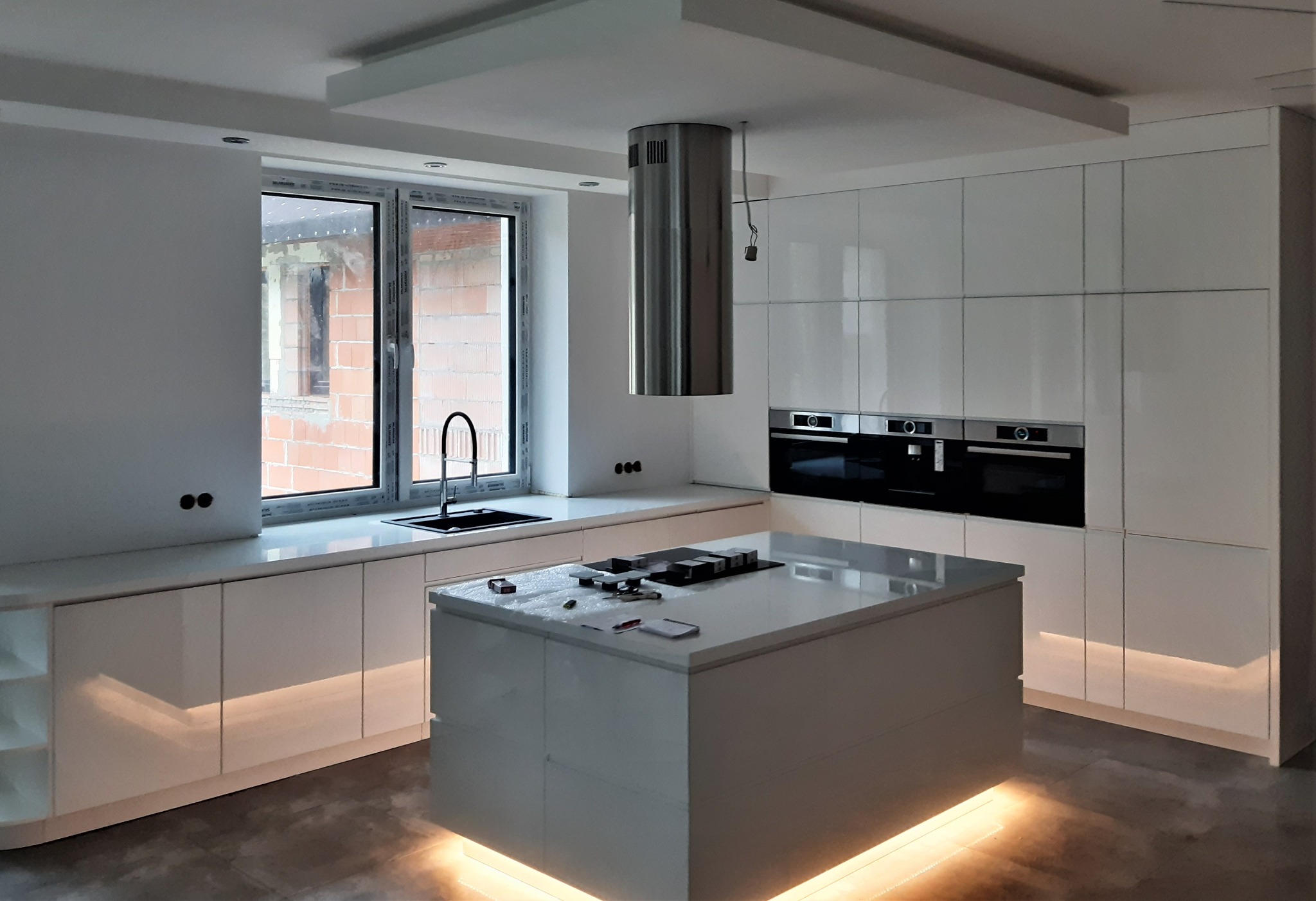 Biała, nowoczesna kuchnia z wyspą i podświetleniem LED, widok na okno z surową ścianą budynku w tle, srebrny okrągły okap nad wyspą.