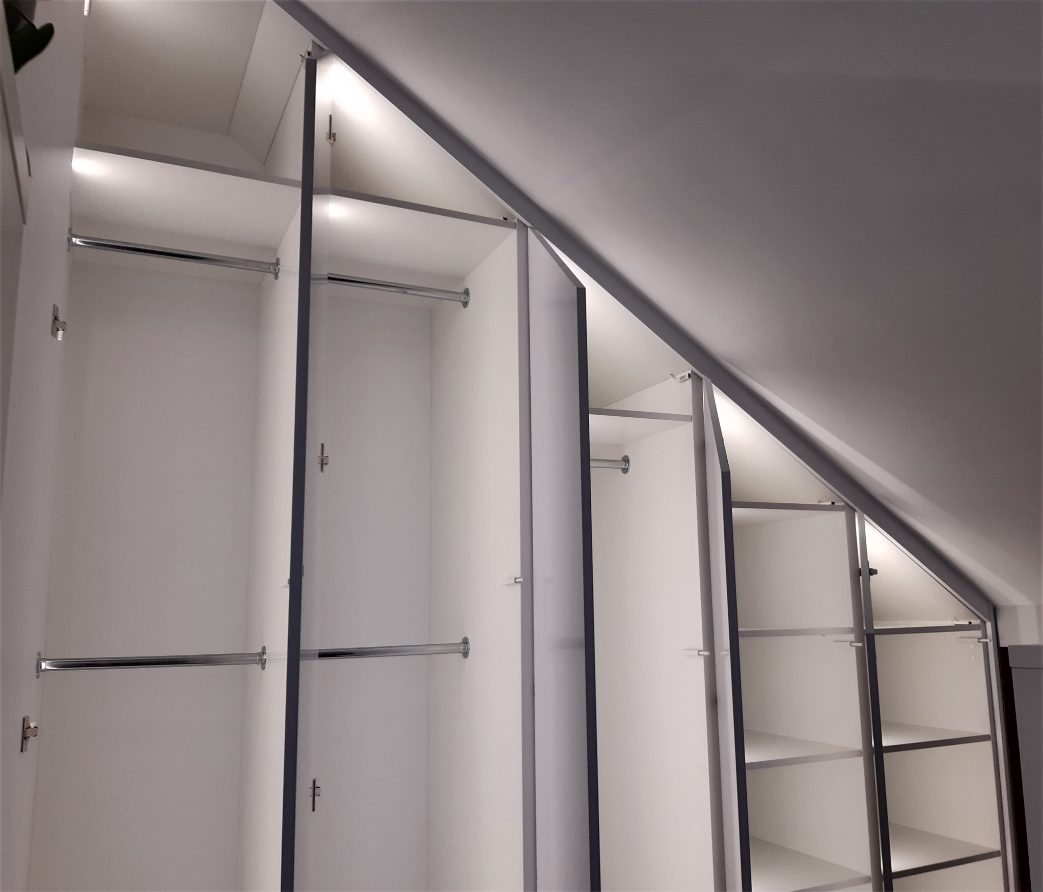 Biała szafa wnękowa z oświetleniem LED, dopasowana do skośnego sufitu na poddaszu, widok od dołu.