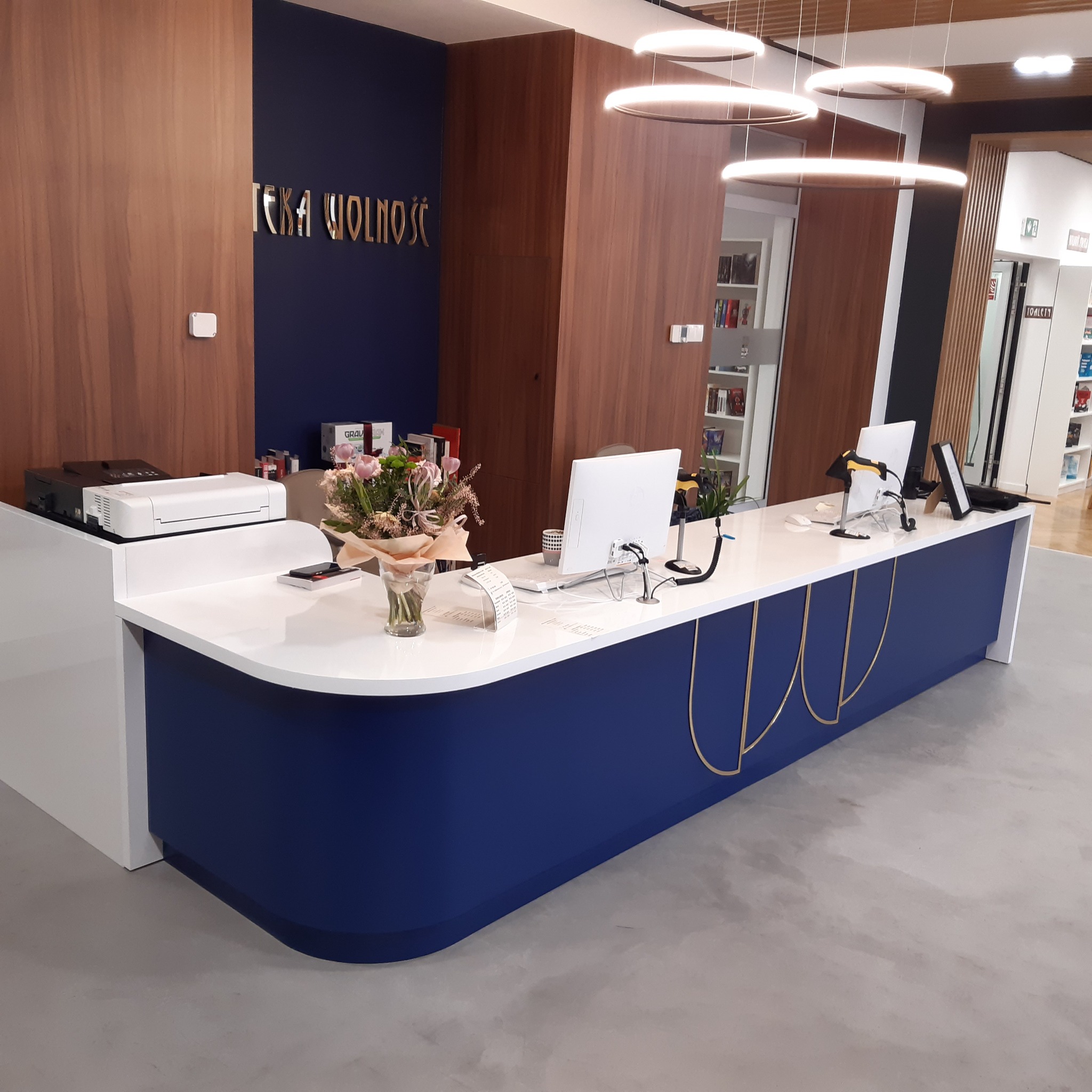 Recepcja w bibliotece z białym blatem, niebieskim frontem zdobionym złotymi łukami, kwiatami i komputerami.