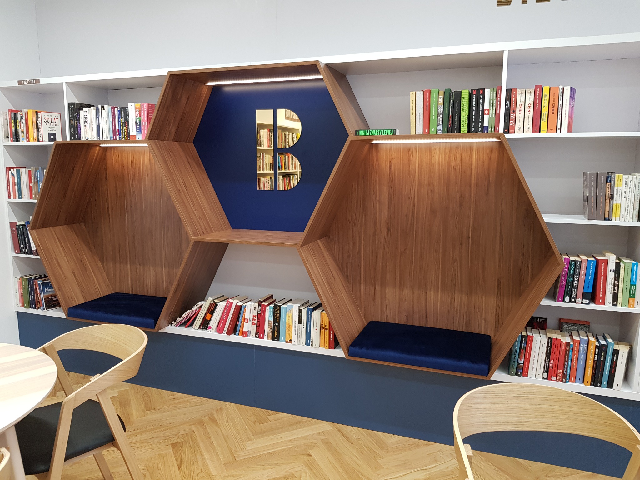 Nowoczesna biblioteczka z drewnianymi wnękami do czytania, podświetlane półki, niebieskie akcenty kolorystyczne i drewniana podłoga w jodełkę.