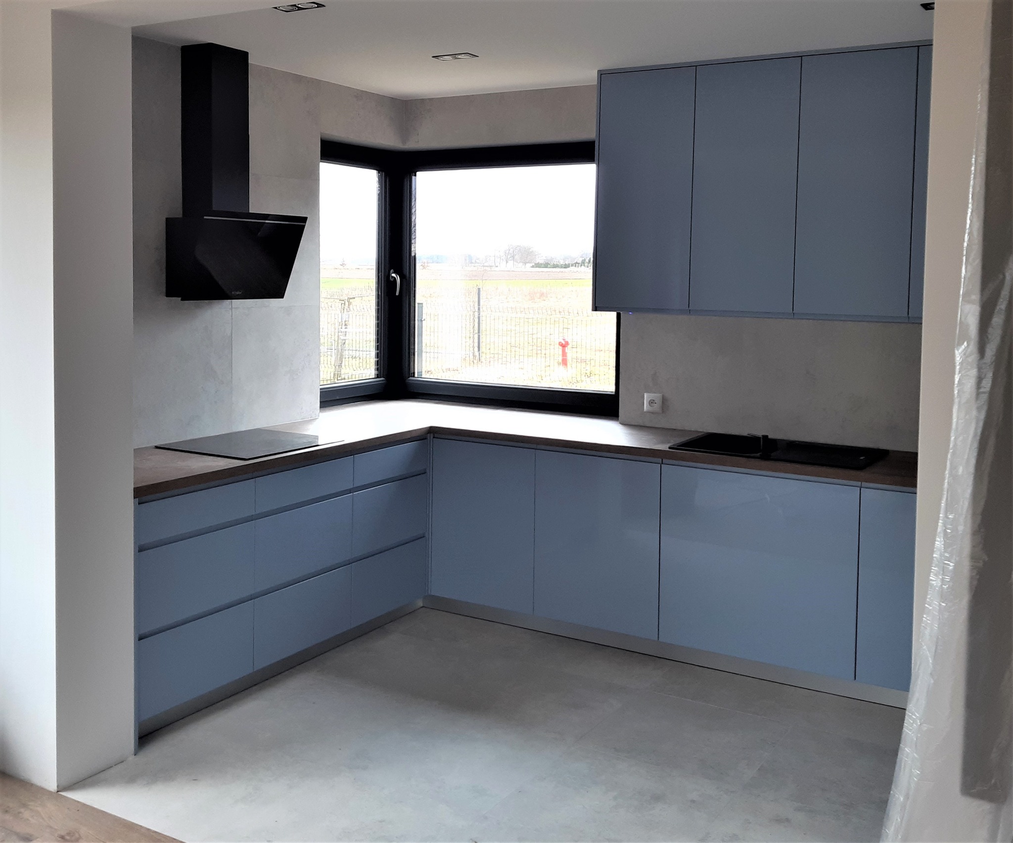 Niebieska, nowoczesna kuchnia z połyskiem, czarny okap i płyta indukcyjna, narożne okno z widokiem na pole, szara ściana i podłoga, widoczne elementy zabezpieczające przed zakończeniem prac.