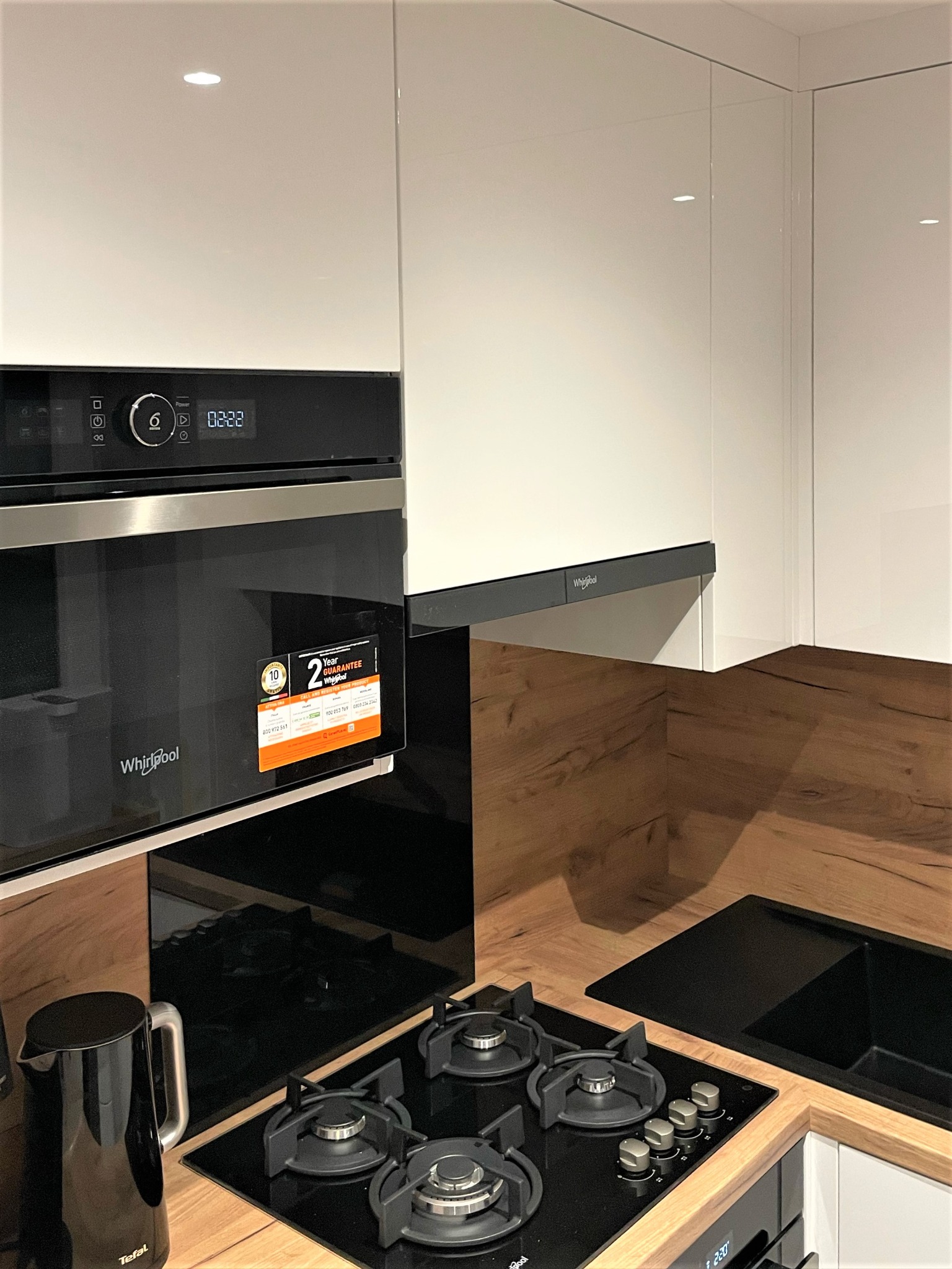 Nowoczesna kuchnia z białymi, lakierowanymi szafkami, drewnianym blatem i czarnym sprzętem AGD: piekarnikiem Whirlpool z naklejką gwarancyjną, płytą gazową i czajnikiem elektrycznym Tefal.