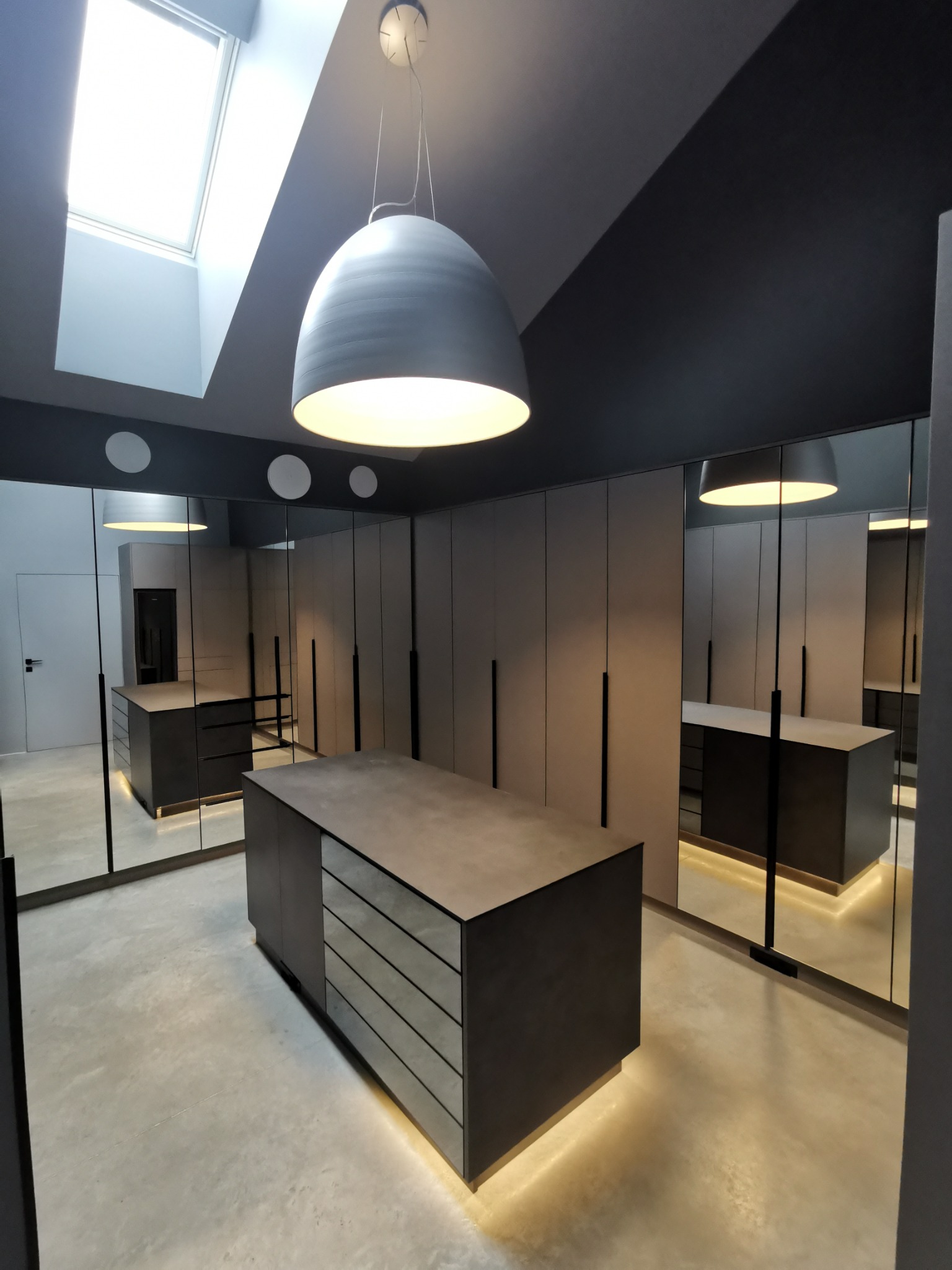Nowoczesna garderoba z szafami wnękowymi z szarymi frontami, oświetleniem LED przy podłodze i wyspą z szufladami pod sufitem z oknem dachowym.