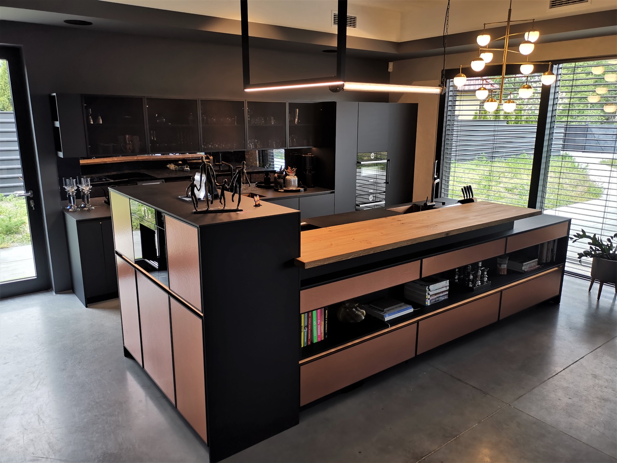 Nowoczesna kuchnia z wyspą, czarne szafki z metalowymi frontami, drewniany blat wyspy, designerskie oświetlenie i dekoracyjne figurki.