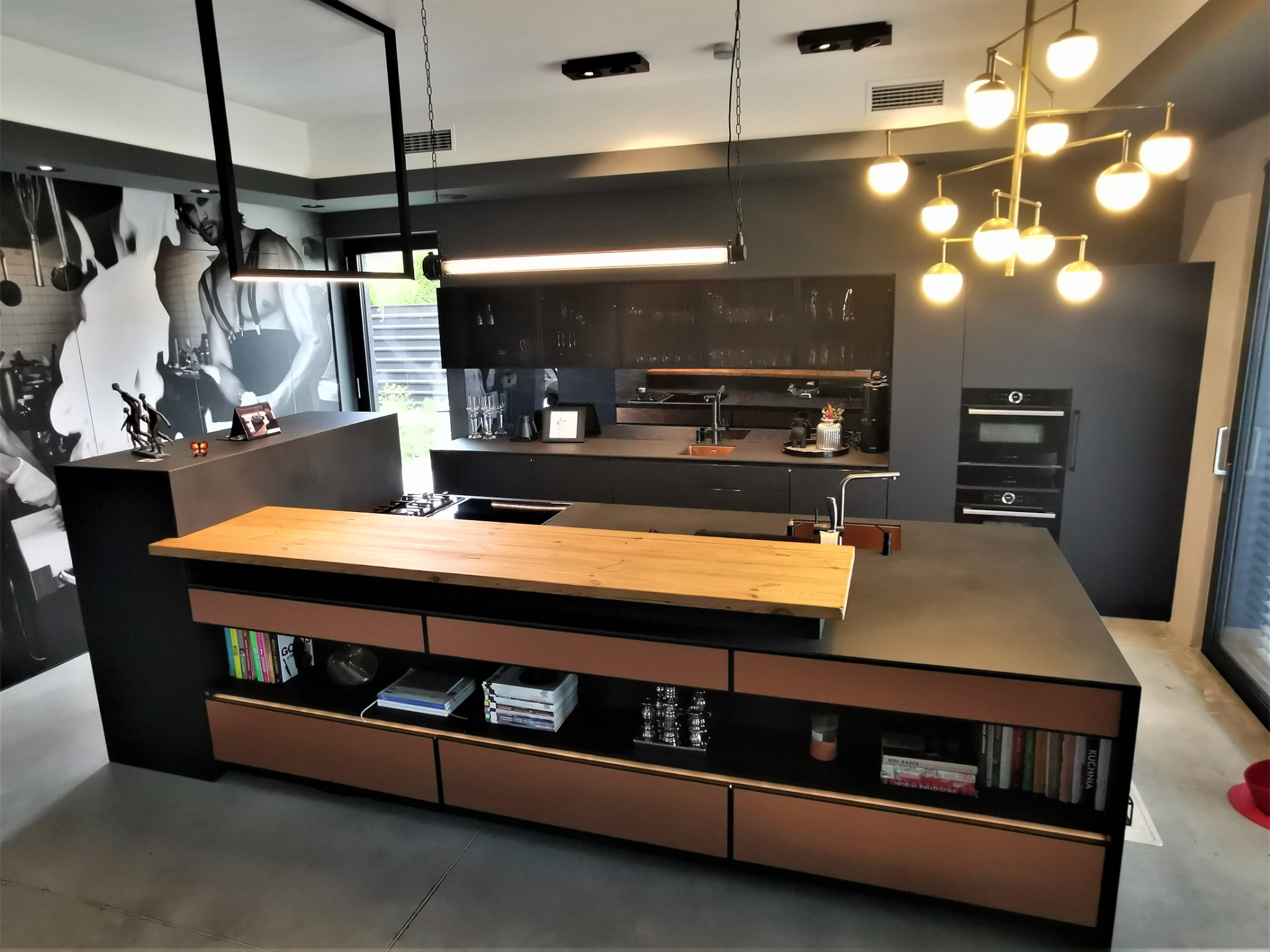 Nowoczesna kuchnia w ciemnych kolorach z wyspą z drewnianym blatem, oświetlona designerskimi lampami wiszącymi, z dekoracyjną fototapetą na ścianie.