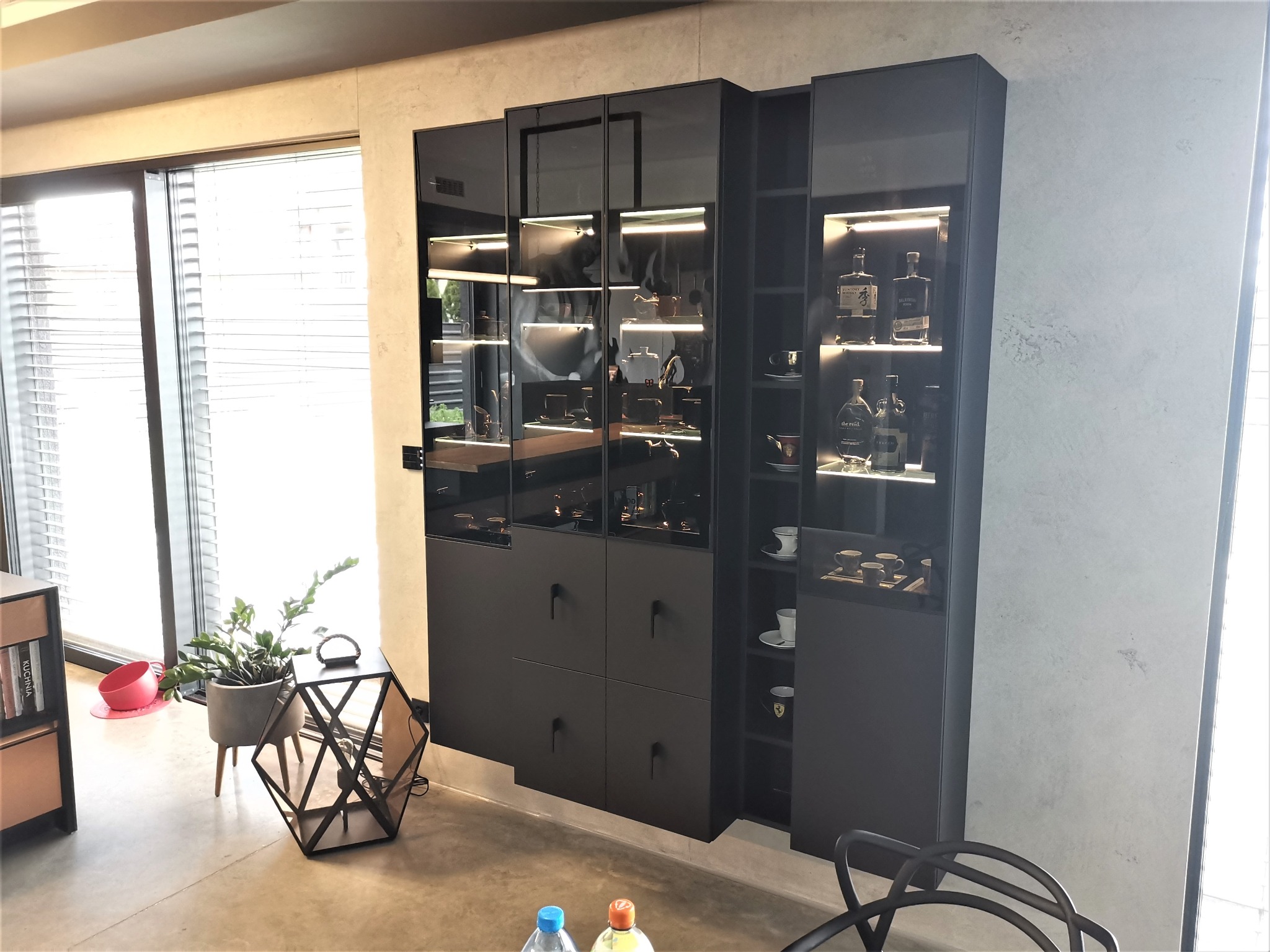 Czarne, nowoczesne meble do przechowywania z przeszklonymi witrynami, podświetlone od wewnątrz, eksponujące szkło i porcelanę w salonie z dużym oknem.