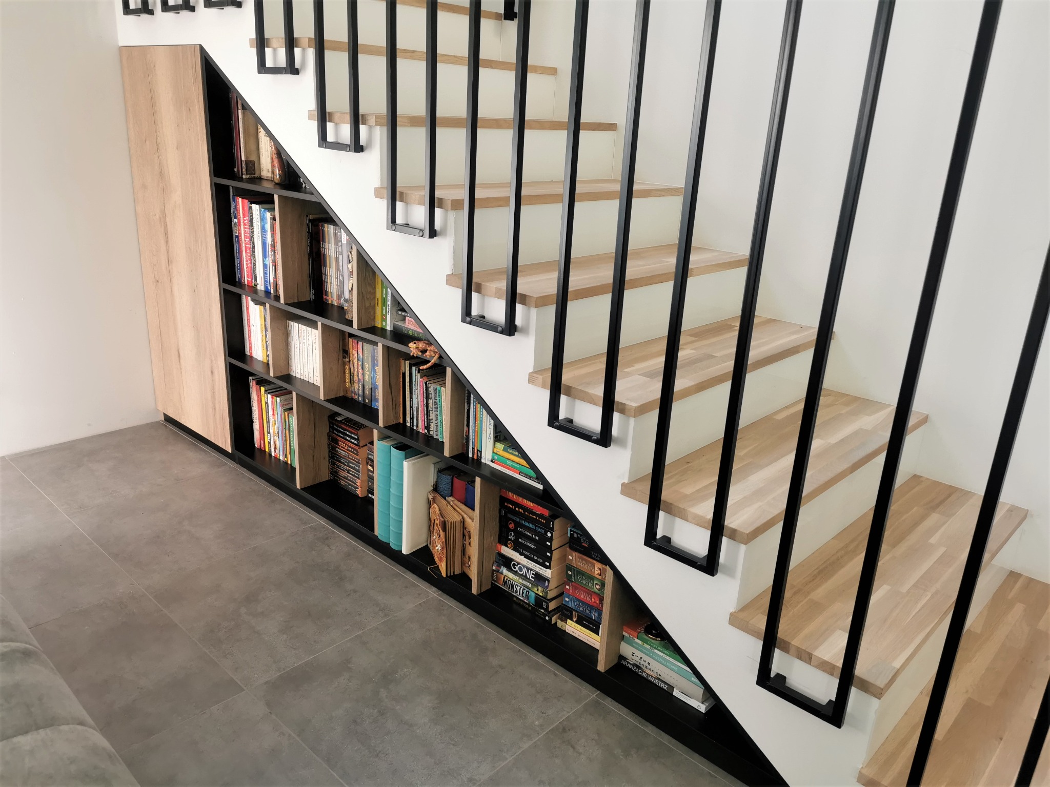 Zabudowa pod schodami z półkami na książki, czarne obramowanie, drewniane schody z czarnymi barierkami, szara podłoga.