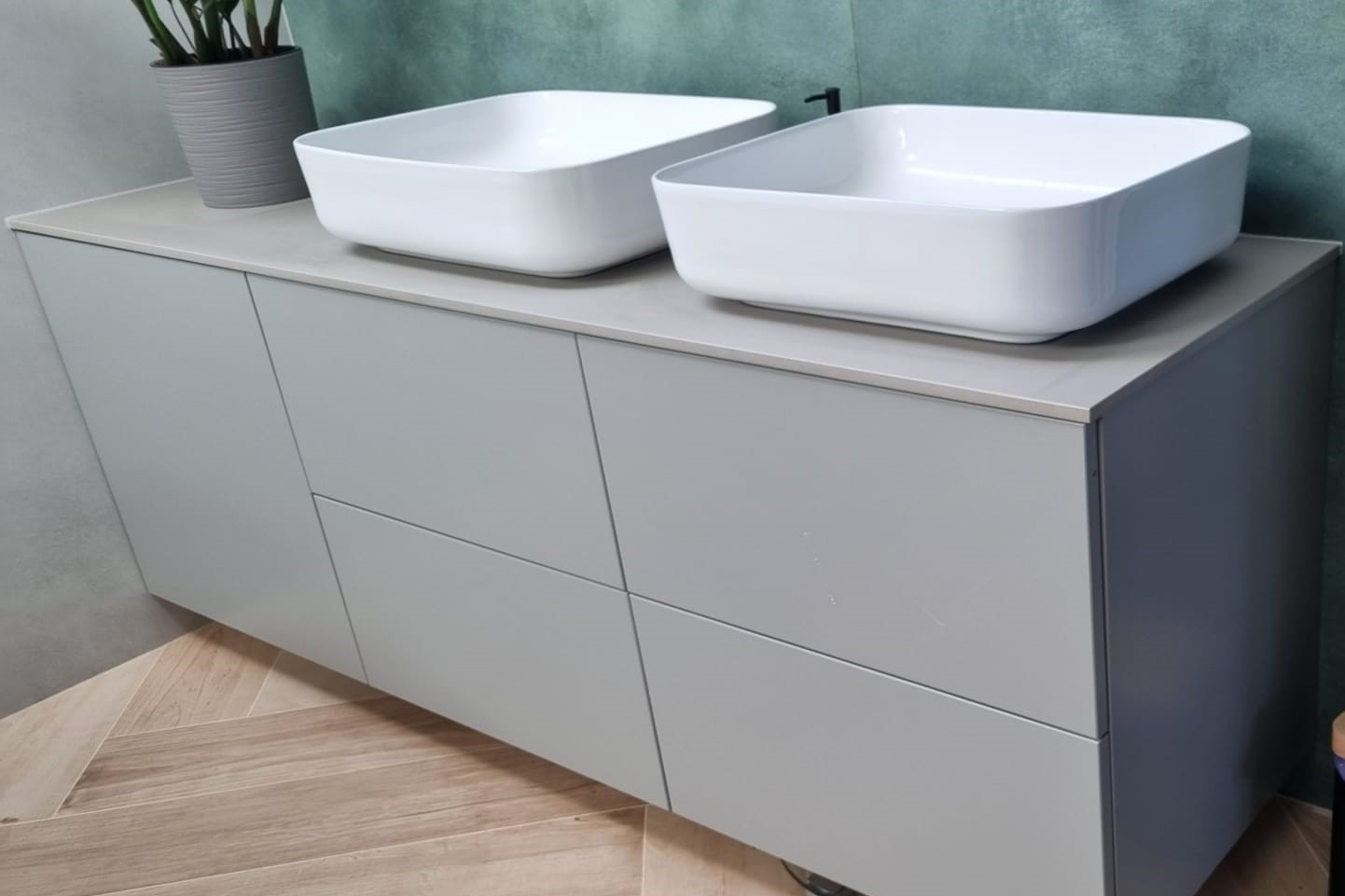 Dwie kwadratowe, białe umywalki nablatowe na szarej szafce z czterema frontami, na podłodze parkiet ułożony we wzór jodełki.