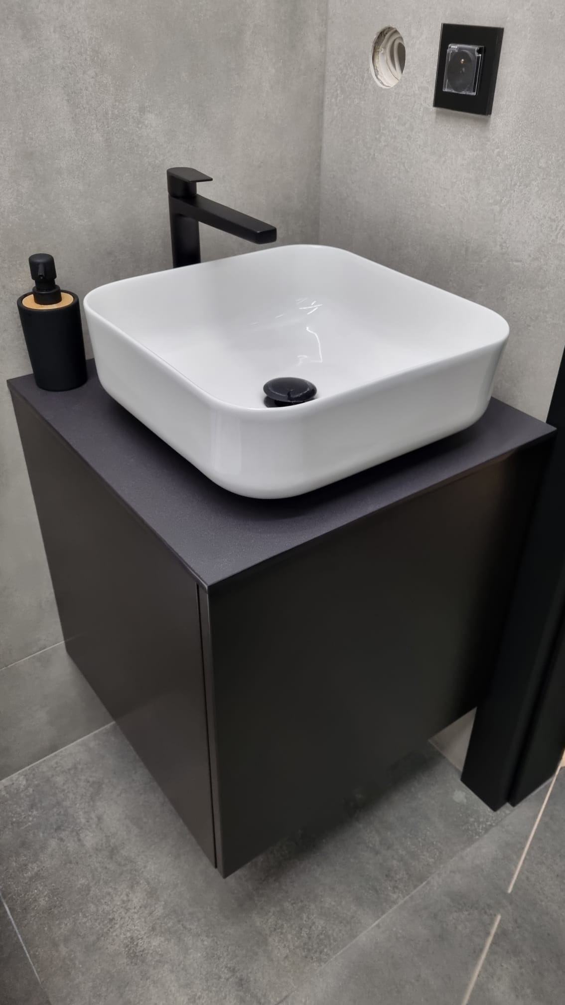Biała, kwadratowa umywalka nablatowa z czarną armaturą i dozownikiem mydła na ciemnoszarej szafce w nowoczesnej łazience z szarymi płytkami.