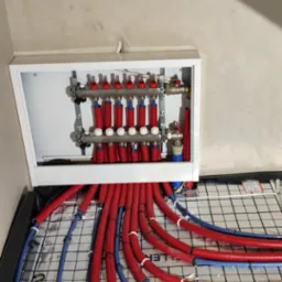 Rozdzielacz ogrzewania podłogowego w białej obudowie, z widocznymi czerwonymi rurami grzewczymi i niebieskimi rurami doprowadzającymi, ułożonymi na siatce montażowej.