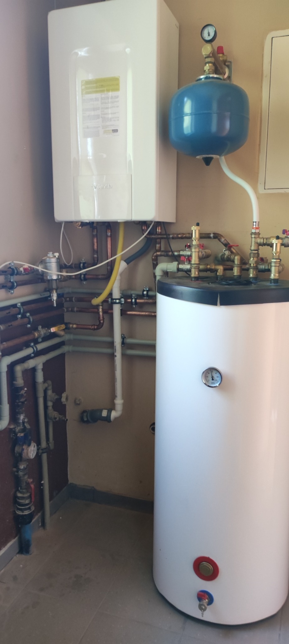 Biały piec gazowy marki Beretta, z niebieskim zbiornikiem wyrównawczym i białym zasobnikiem ciepłej wody, połączone miedzianymi rurami w kotłowni.