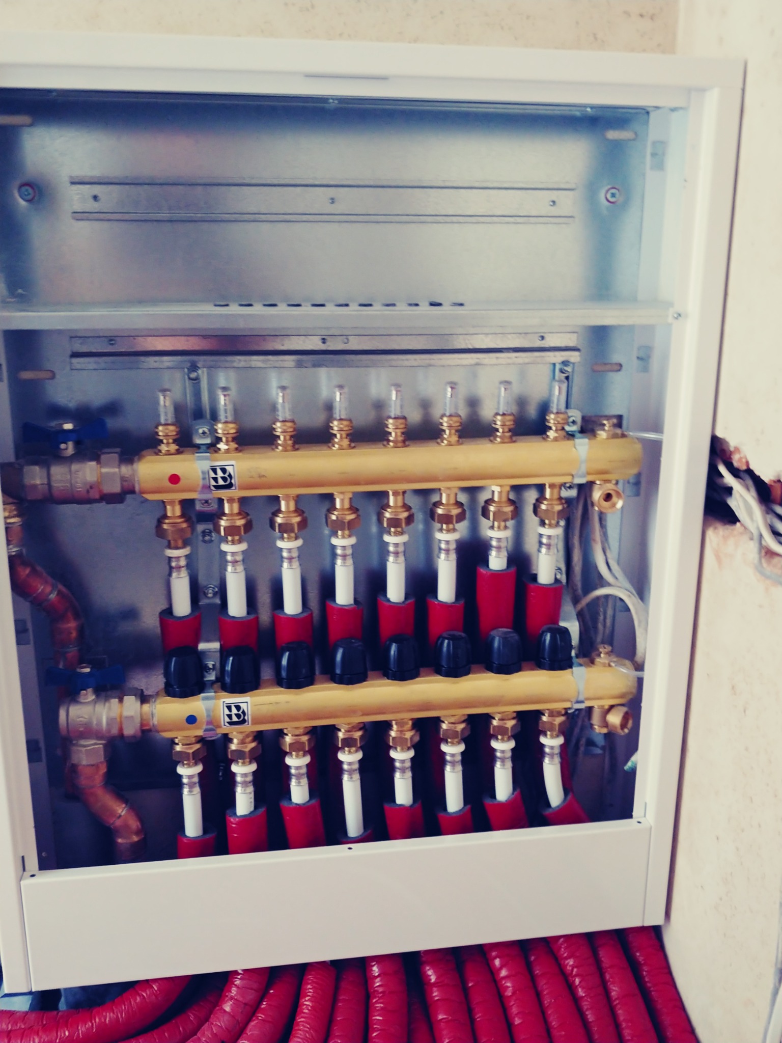 Rozdzielacz ogrzewania podłogowego w białej szafce, widoczne czerwone rury w izolacji, mosiężne złączki i zawory regulacyjne.