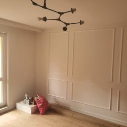 Tomasz Dolacki - Pomalowane na biało ściany z dekoracyjnymi listwami w nowym mieszkaniu, widoczna nowoczesna czarna lampa sufitowa, podłoga wyłożona panelami, karton i worek z odpadami po remoncie przy ścianie.