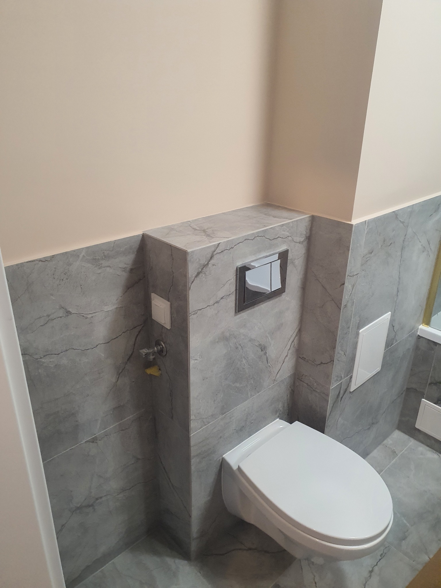 Nowoczesna toaleta podwieszana z szarą marmurową okładziną ścienną, ukryty zbiornik z chromowanym panelem spłukującym i dostępem rewizyjnym.