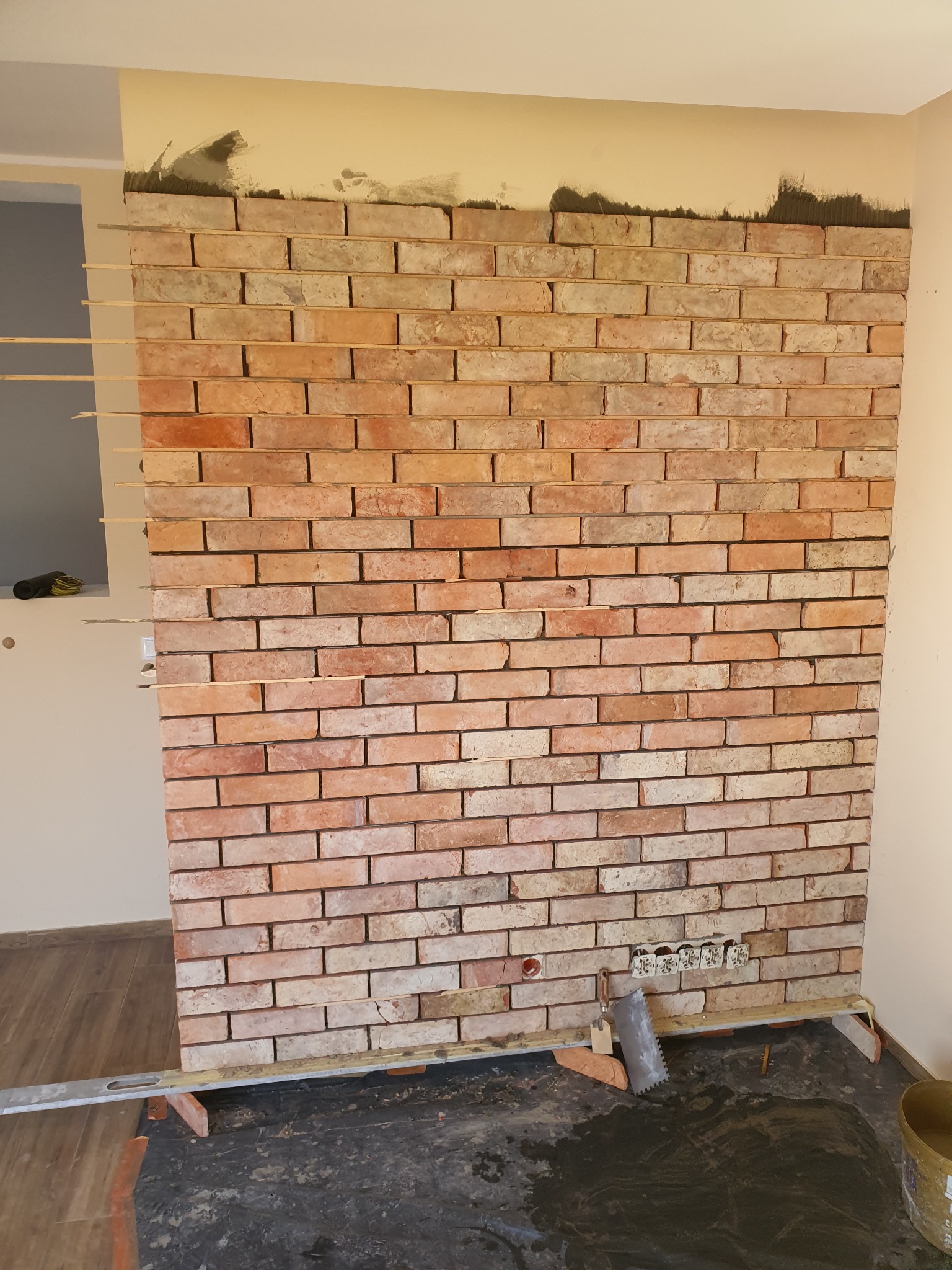 Ściana z cegły klinkierowej w trakcie montażu, widoczne listewki dystansowe, poziomica, puszki elektryczne i narzędzia glazurnicze.