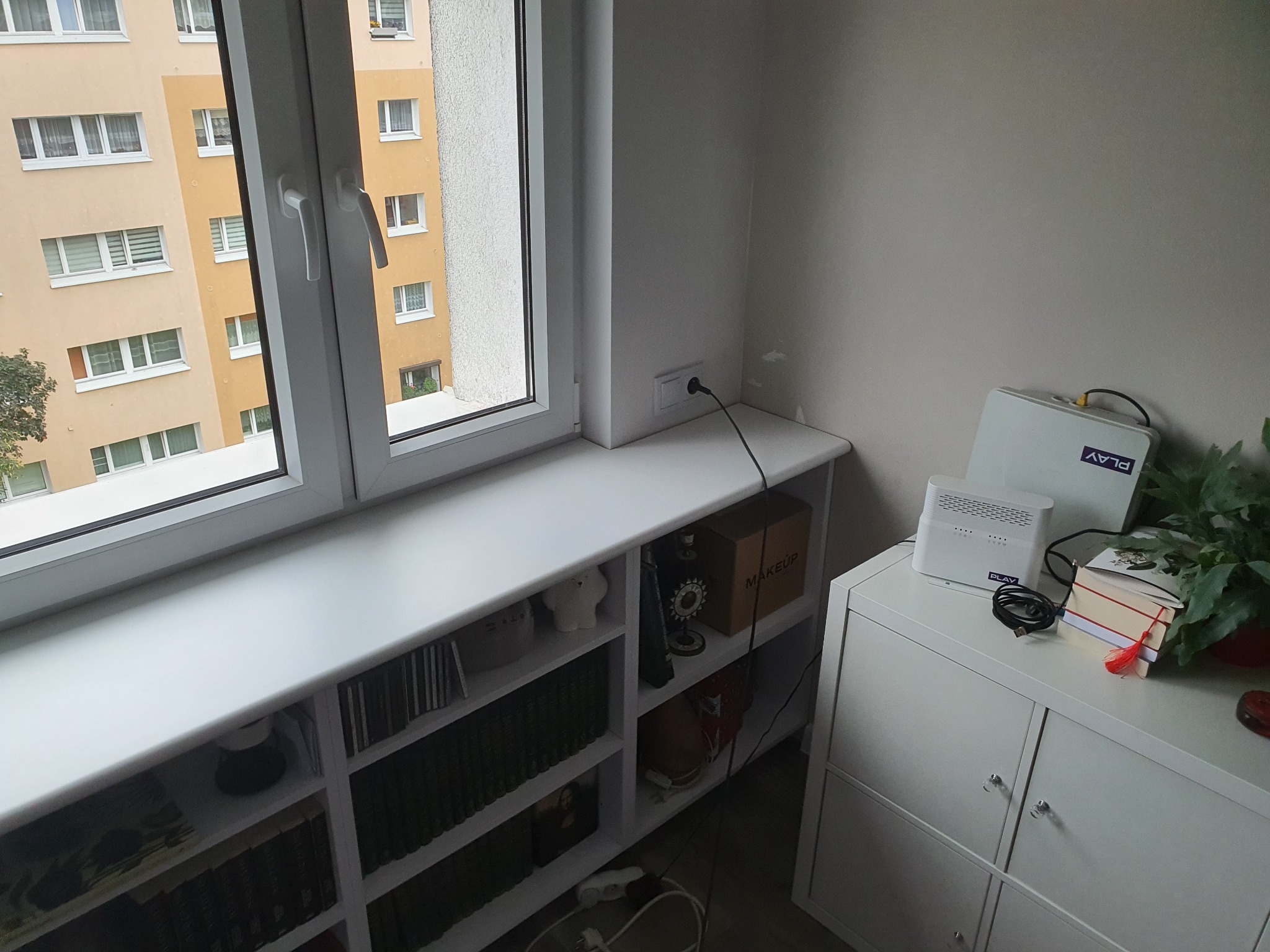 Biały parapet okienny z wbudowanym regałem na książki i dekoracje, obok biała komoda z routerem i rośliną, widok na blok mieszkalny za oknem.