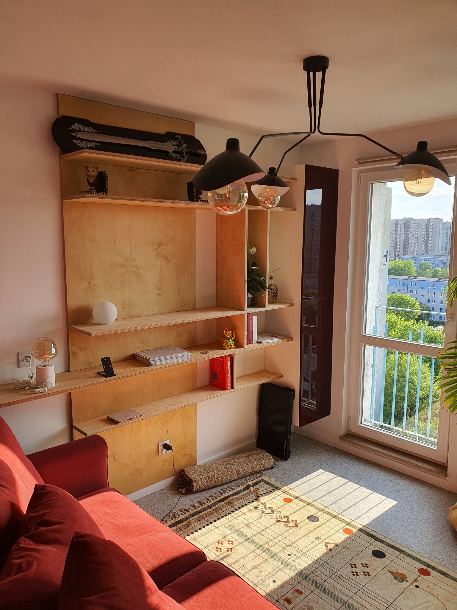 Półki z surowego drewna z dekoracjami w pokoju dziennym w Poznaniu, oświetlone nowoczesną lampą wiszącą, obok czerwona sofa i okno z widokiem na miasto.