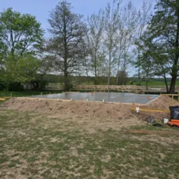 Wylany fundament pod budowę na działce z zielenią i drzewami. Widoczne szalunki, wystające rury. Na pierwszym planie trawa i ziemia.