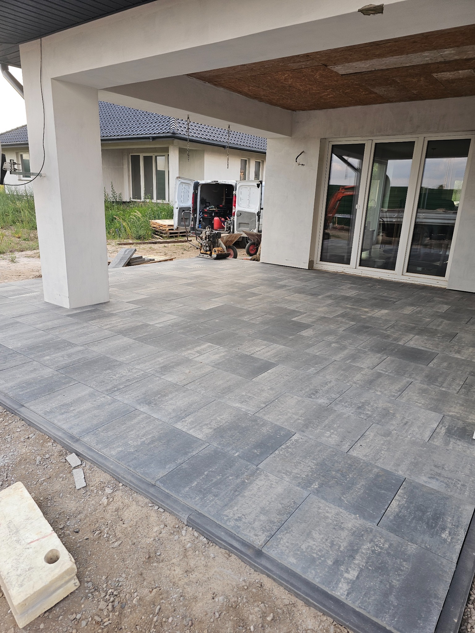 Ułożona szara kostka brukowa na tarasie z widocznym budynkiem w tle i rozładowanym sprzętem budowlanym z samochodu dostawczego.
