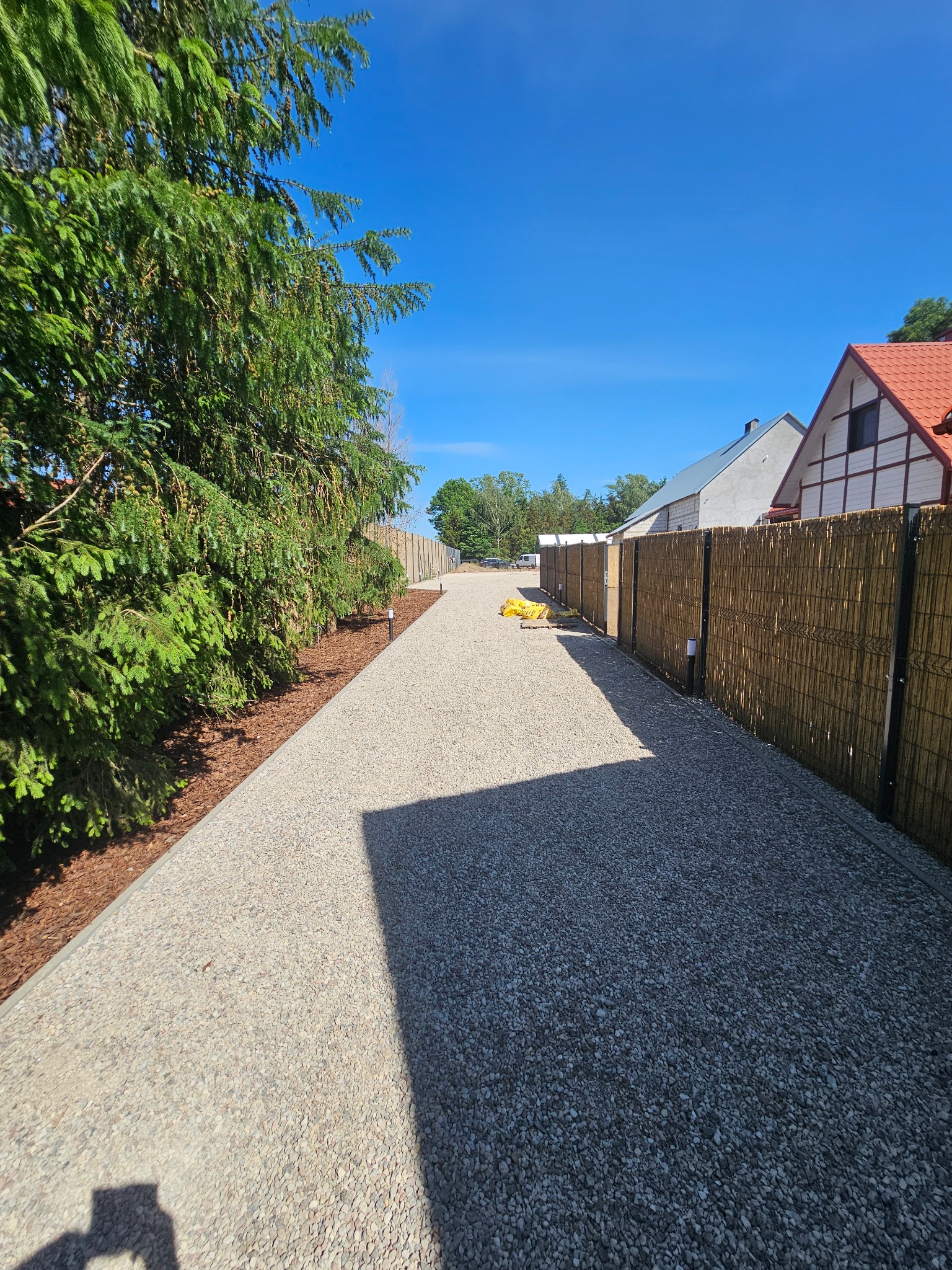 Żwirowa ścieżka prowadząca wzdłuż bambusowego ogrodzenia i drzew, z widocznymi lampkami oświetleniowymi i workami z materiałem budowlanym w tle.