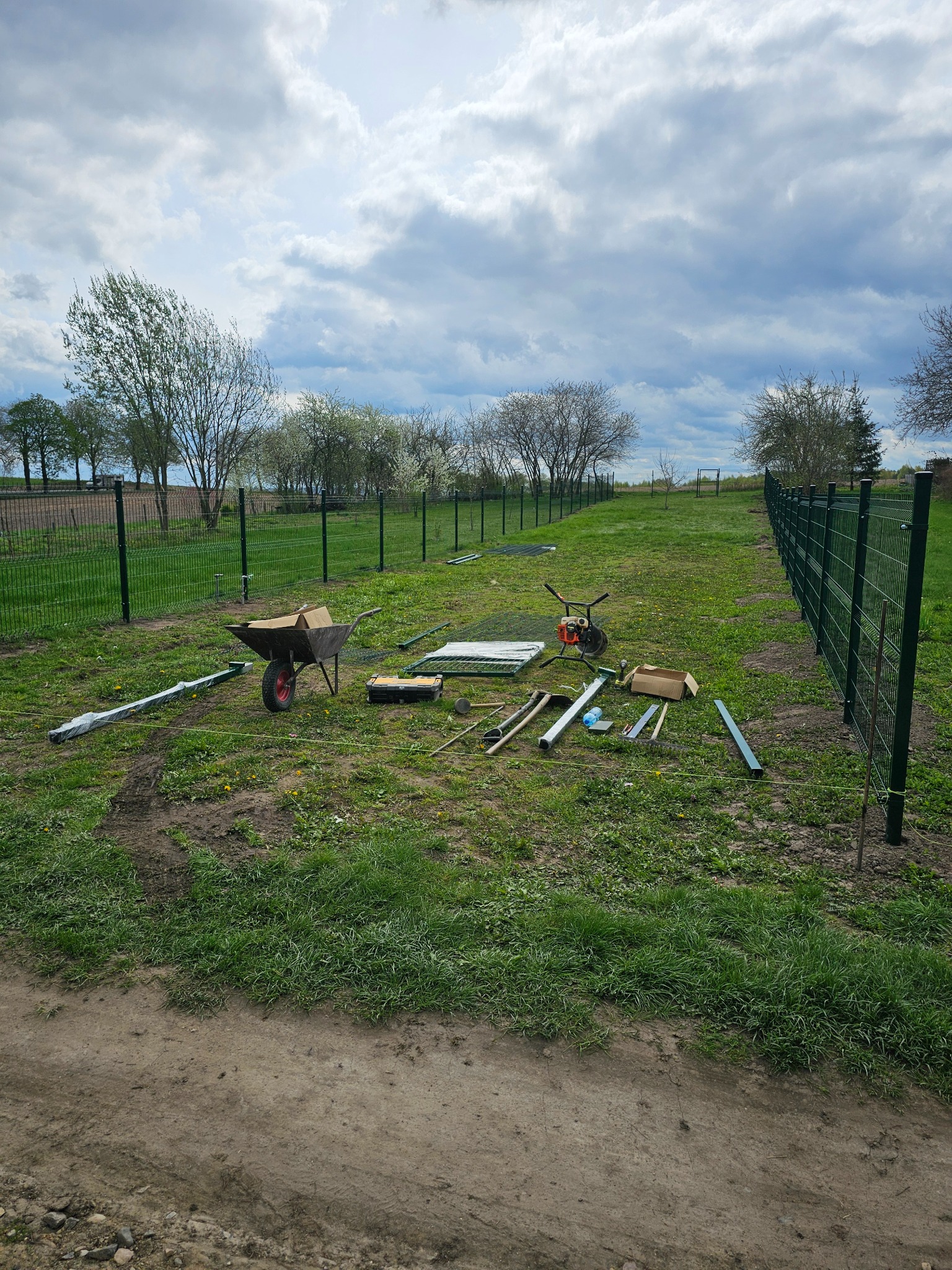 Montaż ogrodzenia panelowego: widok na rozłożone narzędzia, elementy ogrodzenia i częściowo ukończony płot na tle zielonego trawnika i pochmurnego nieba.