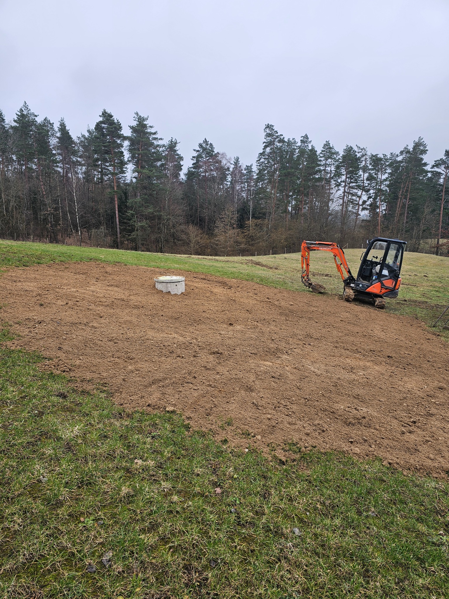 Mini koparka Hitachi na działce z przygotowanym terenem pod instalację, widoczny betonowy element z pokrywą, tło stanowią drzewa.