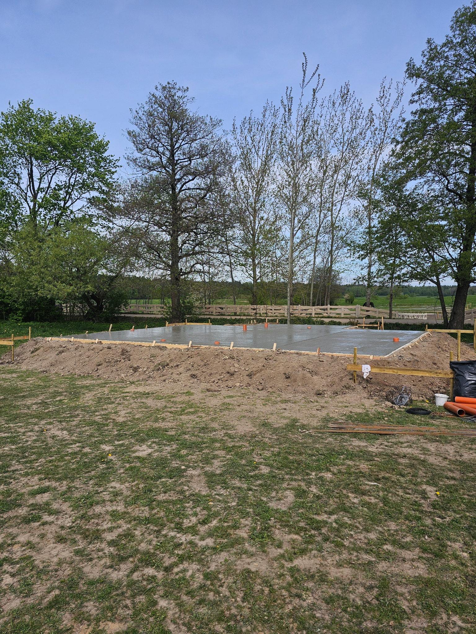 Wylany fundament pod budowę na działce z zielenią i drzewami. Widoczne szalunki, wystające rury. Na pierwszym planie trawa i ziemia.