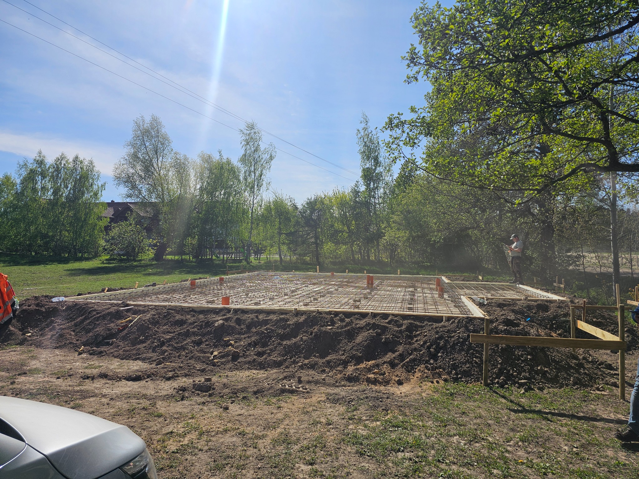 Zbrojenie fundamentu domu jednorodzinnego, widoczne pręty zbrojeniowe i szalunek, w tle drzewa i błękitne niebo.