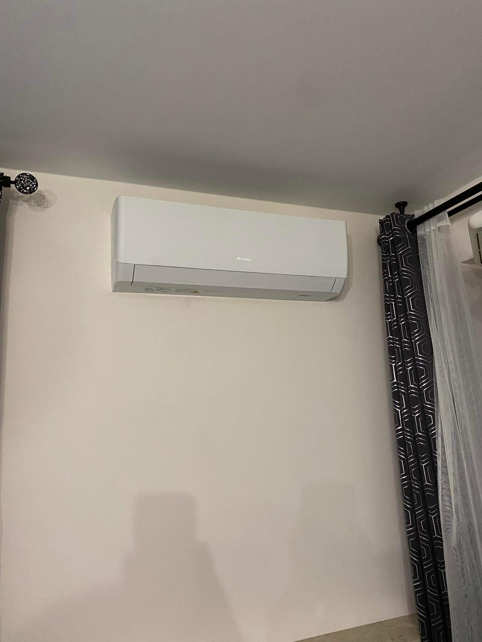 Biała klimatyzacja marki Gree zamontowana na jasnej ścianie w pomieszczeniu z ciemnymi zasłonami w geometryczne wzory i białymi firankami.