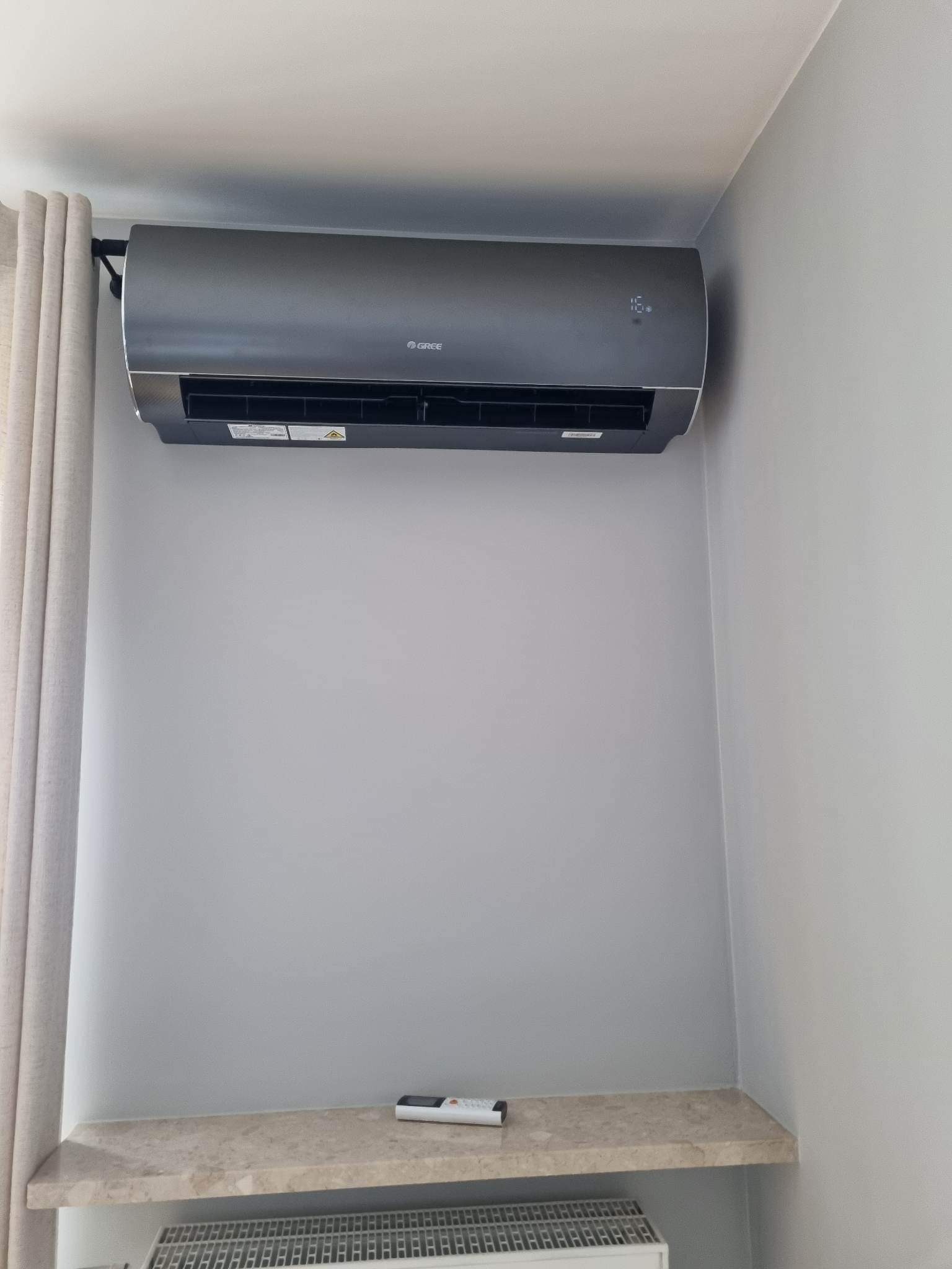 Szary klimatyzator marki Gree zamontowany na ścianie w pomieszczeniu w Milanowie, widoczny pilot na półce poniżej.