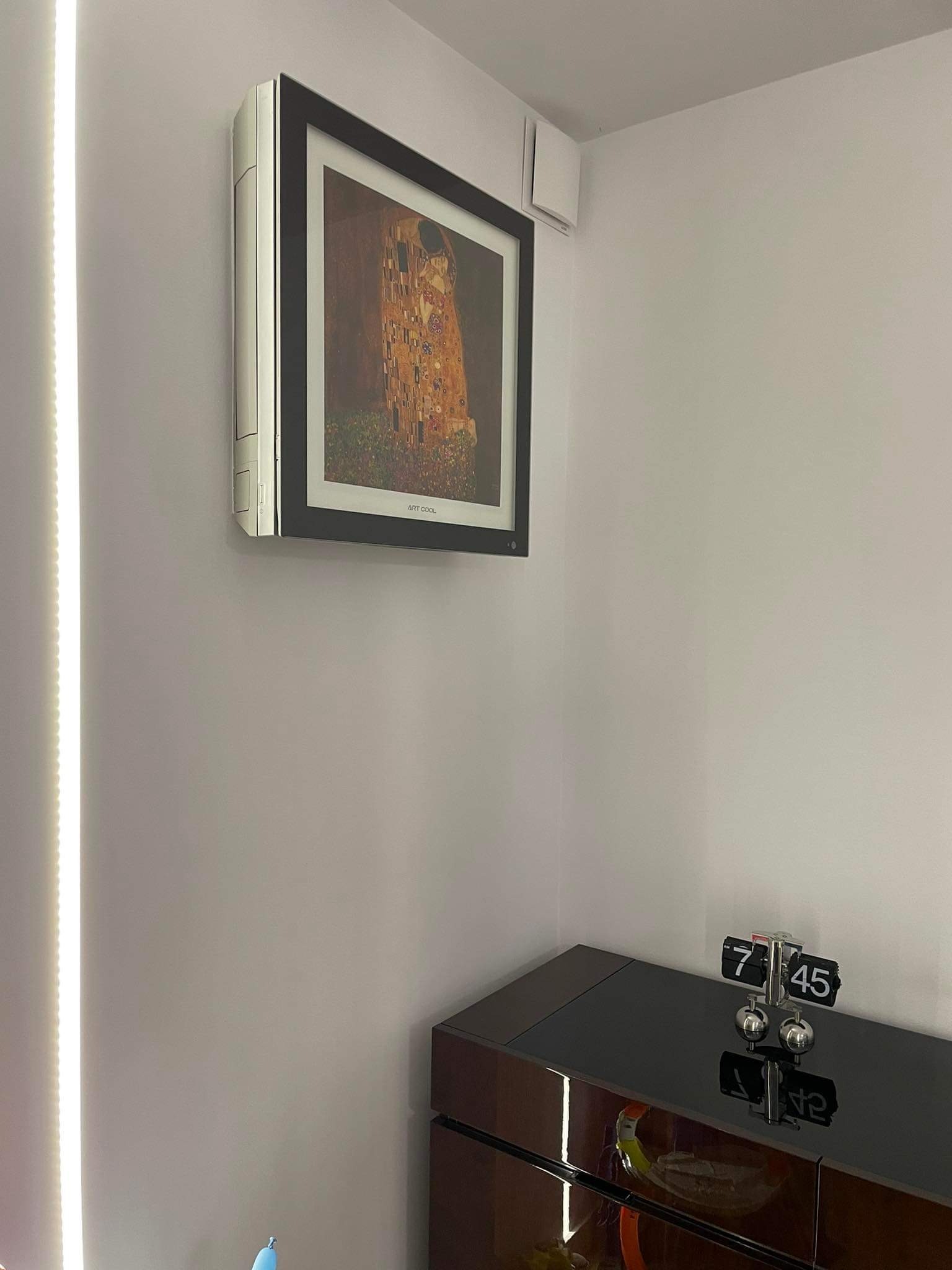 Nowoczesny klimatyzator ścienny z funkcją wyświetlania obrazu, prezentujący reprodukcję 'Pocałunku' Gustava Klimta, zamontowany w minimalistycznym wnętrzu nad błyszczącą komodą z zegarem przerzutowym.