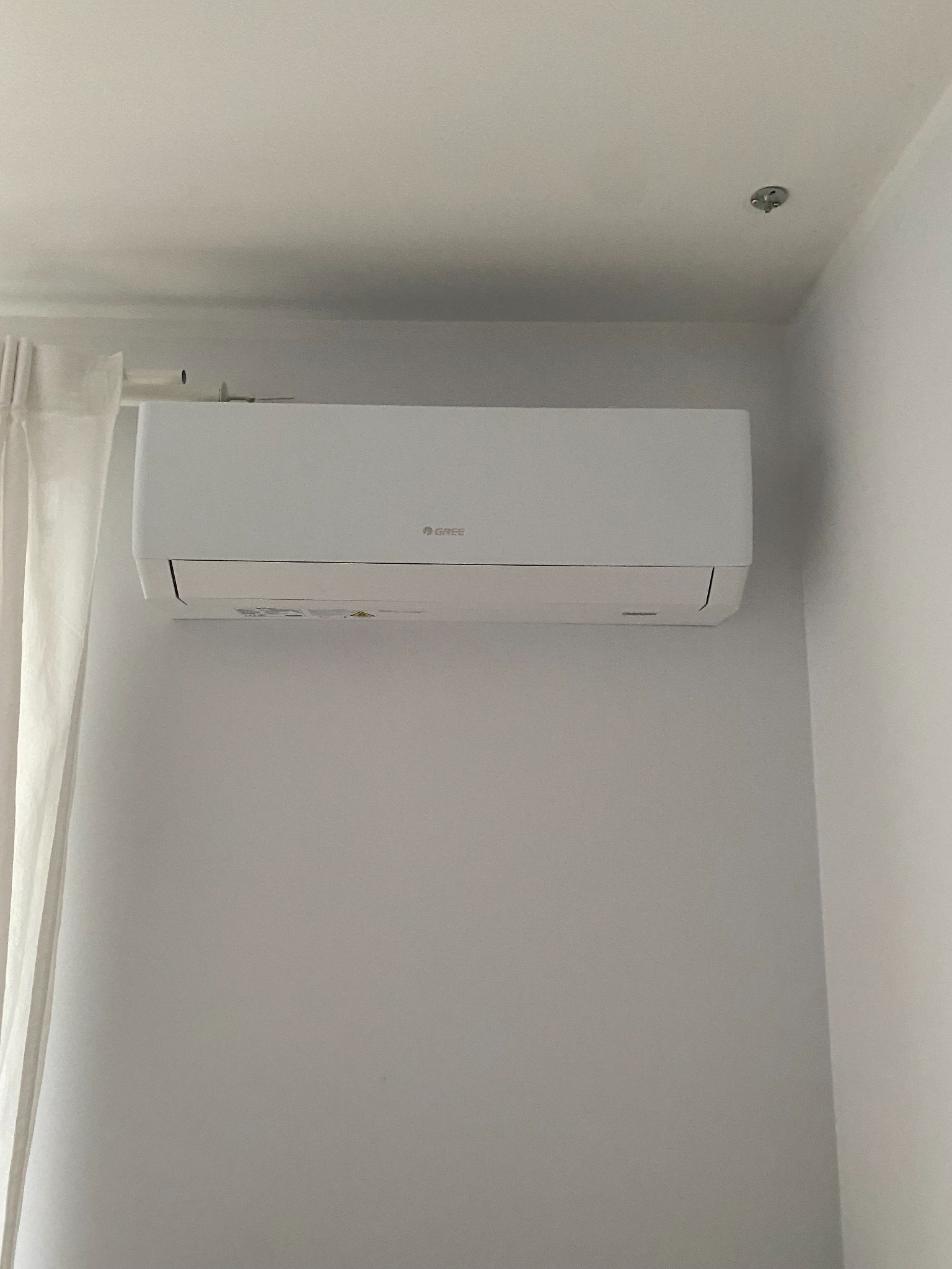 Biały klimatyzator marki Gree zamontowany na białej ścianie w jasnym pomieszczeniu, widoczna zasłona i punkt oświetleniowy na suficie.